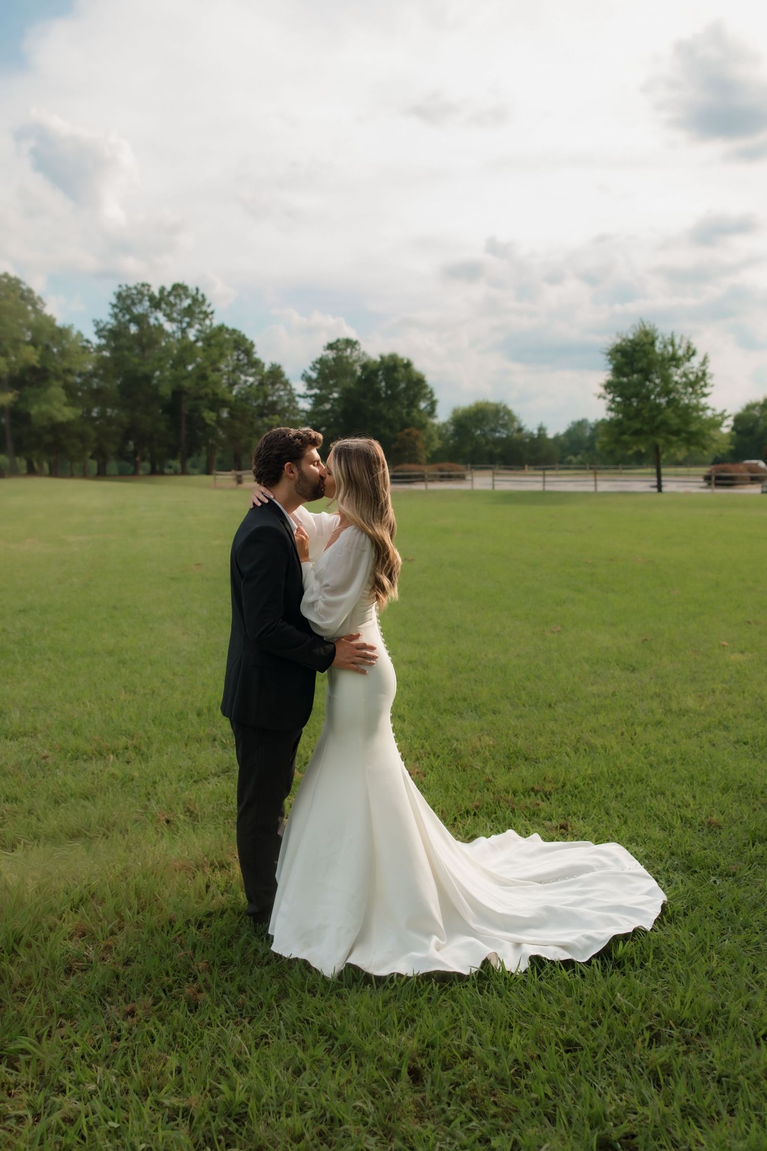 Bride and groom share a romantic kiss in an open green field, bride wearing elegant mermaid wedding gown with train.