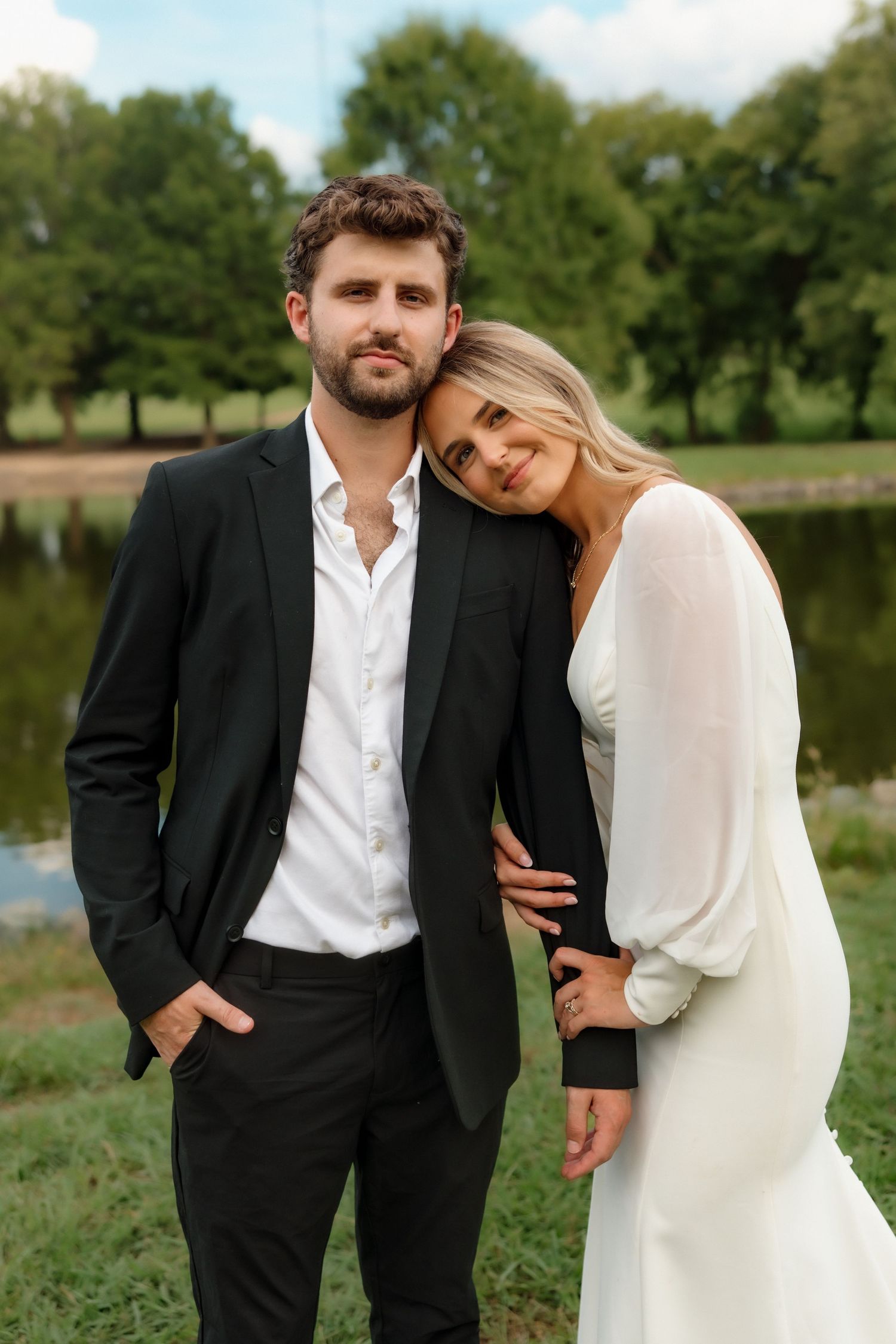 Elegant bride and groom pose together near a lake, bride in white gown leaning on groom.