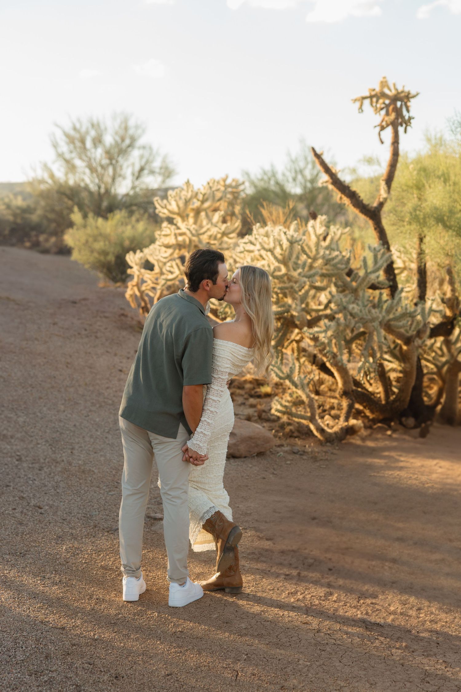 Couple kissing in desert landscape surrounded by cholla cacti on a sandy path during golden hour.