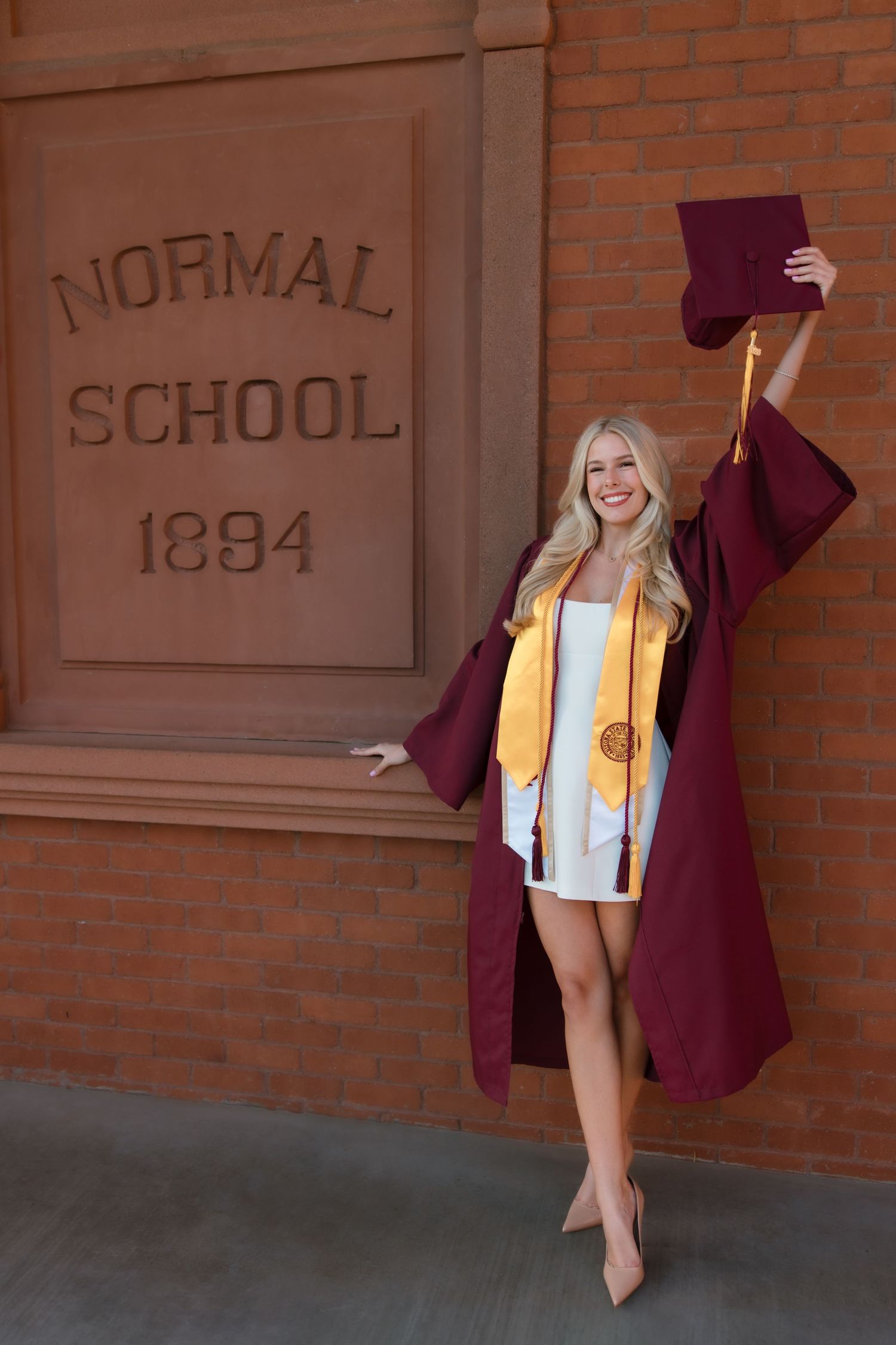 Smiling graduate in maroon cap and gown poses by Normal School 1894 brick wall, holding cap triumphantly.