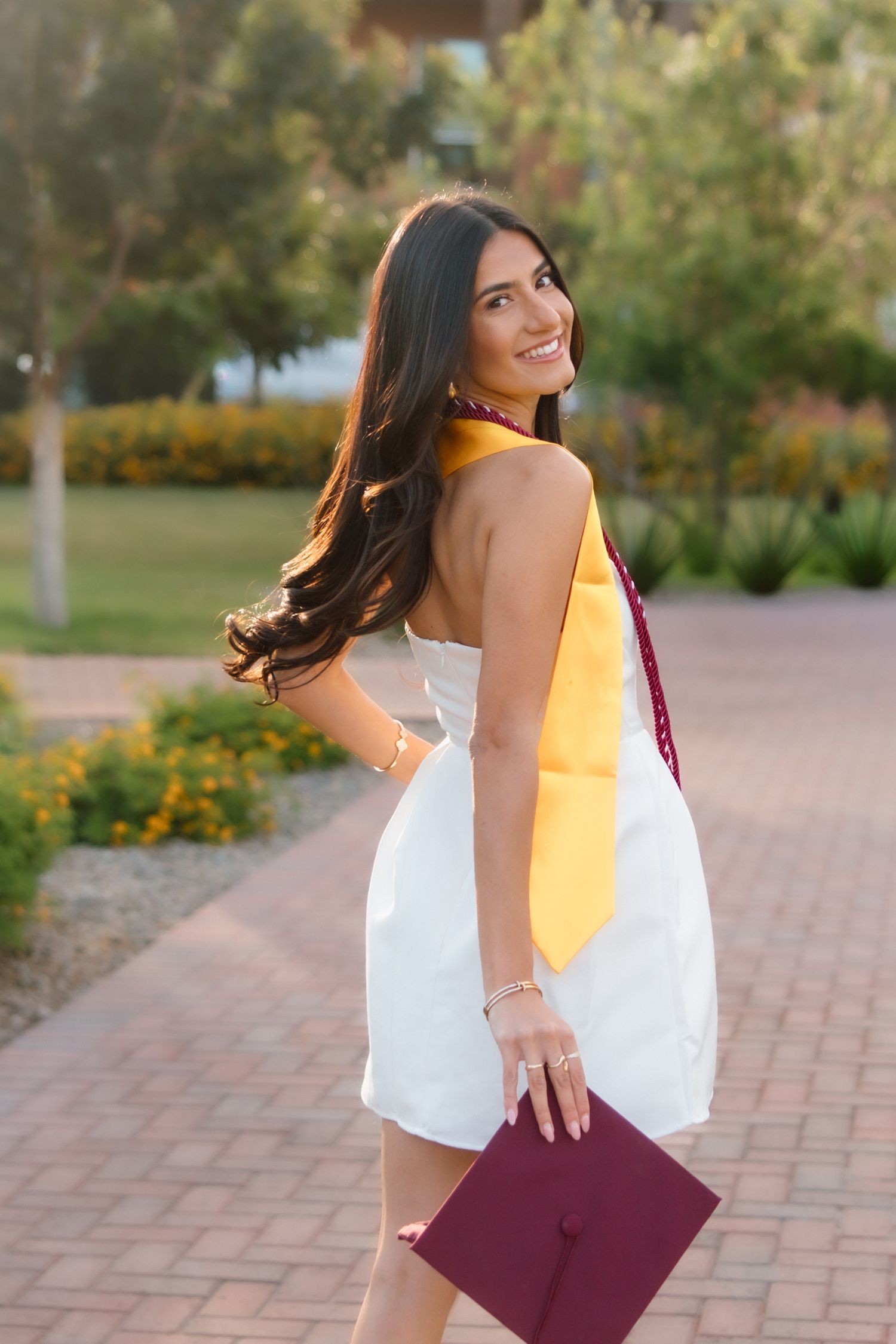 Smiling young woman in white dress holding maroon graduation cap with gold sash outdoors on a sunny day.