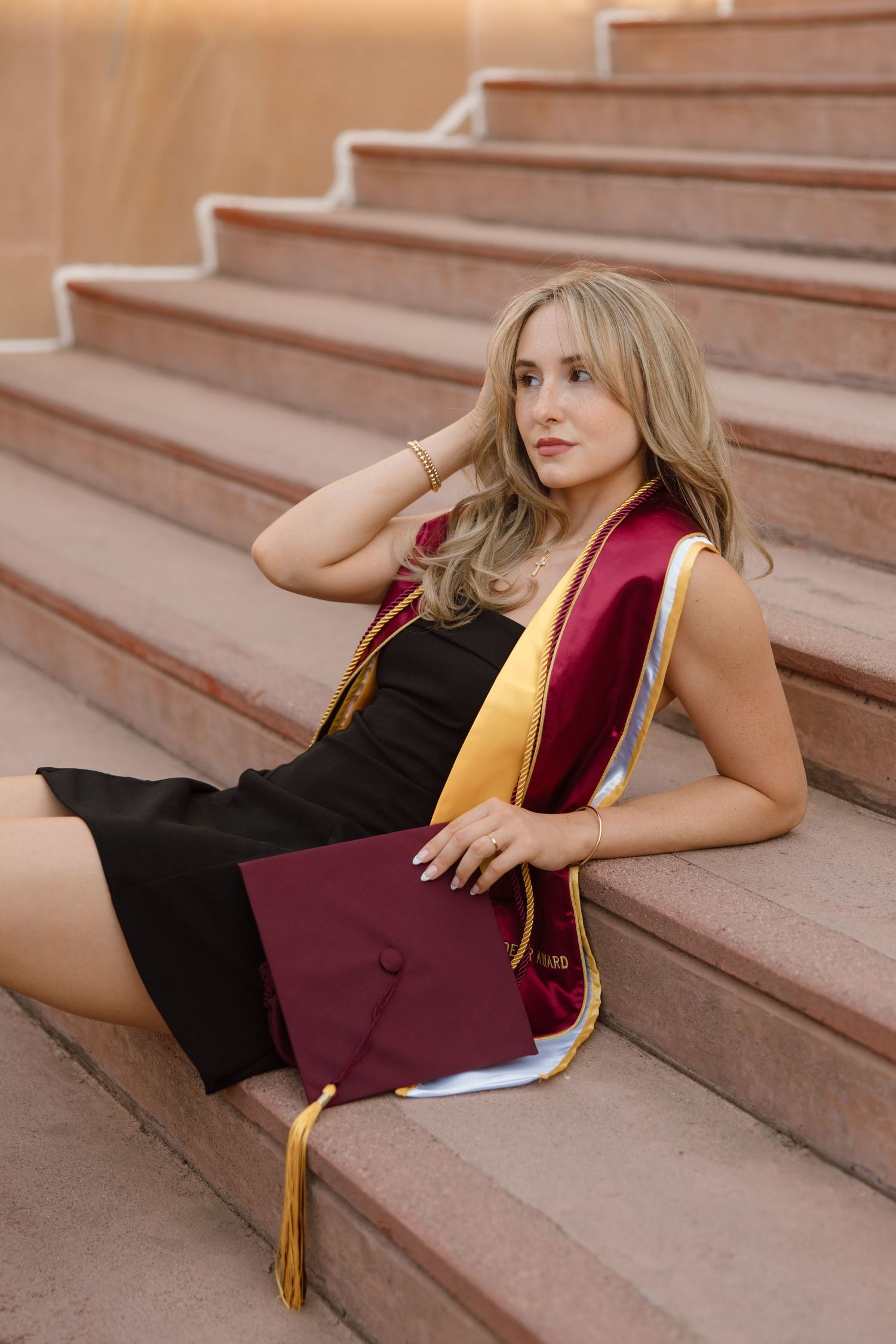 Blonde woman in black dress with maroon and gold graduation regalia sits on stone steps holding a cap.
