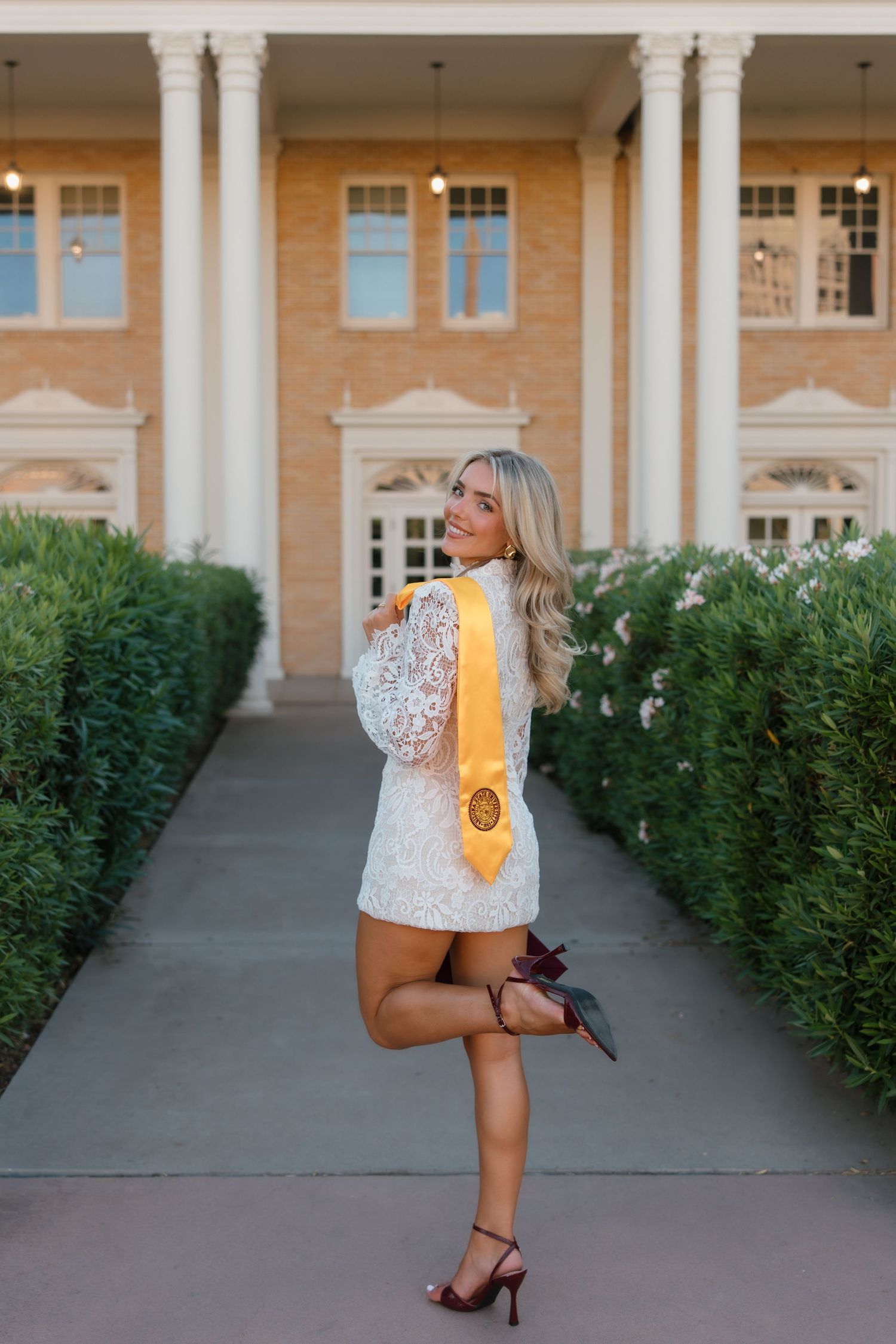 Blonde woman in white lace dress with gold graduation sash posing in front of a brick building.