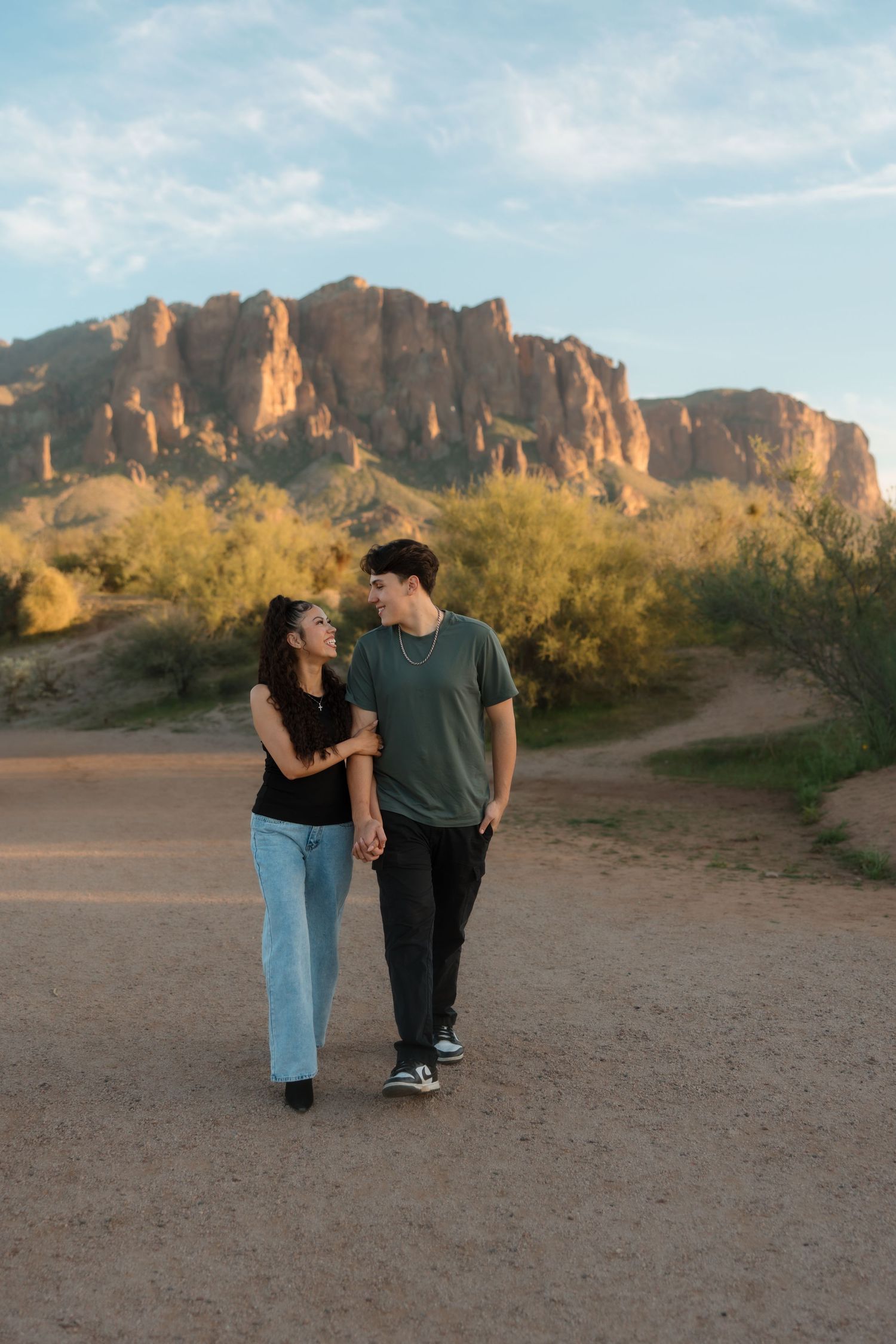 A couple walks hand in hand on a desert trail with dramatic rocky mountains in the background at dusk.