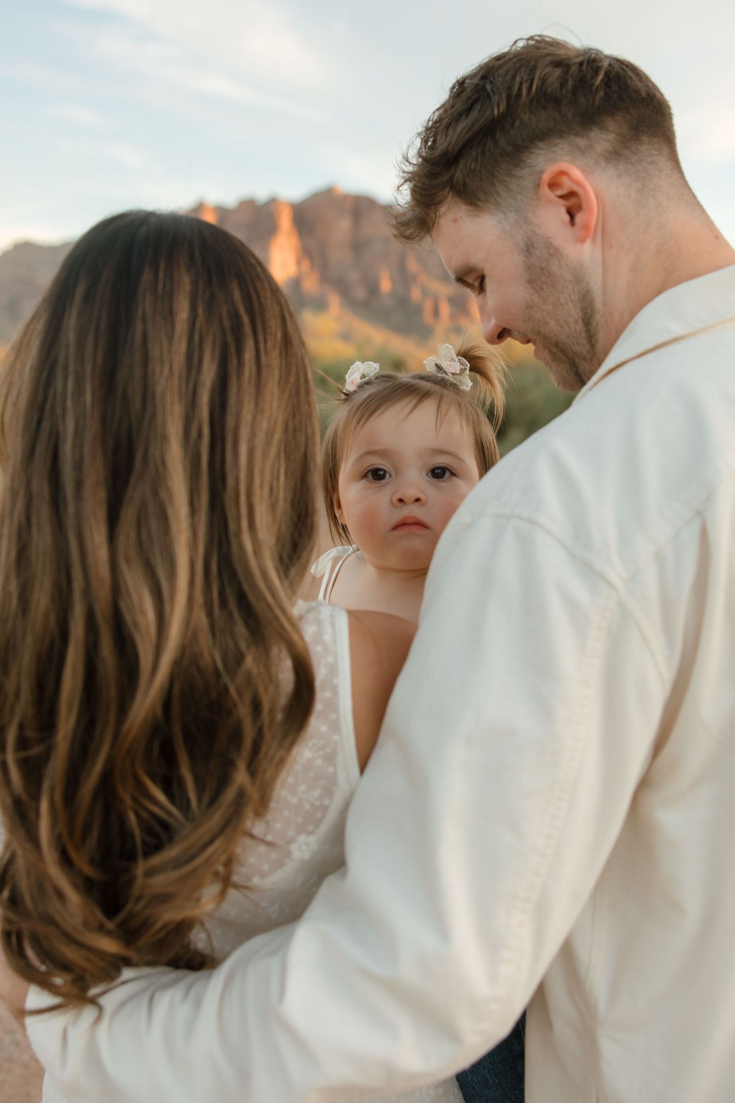 Toddler girl with flower headband looking at camera while being held by parents in desert setting.