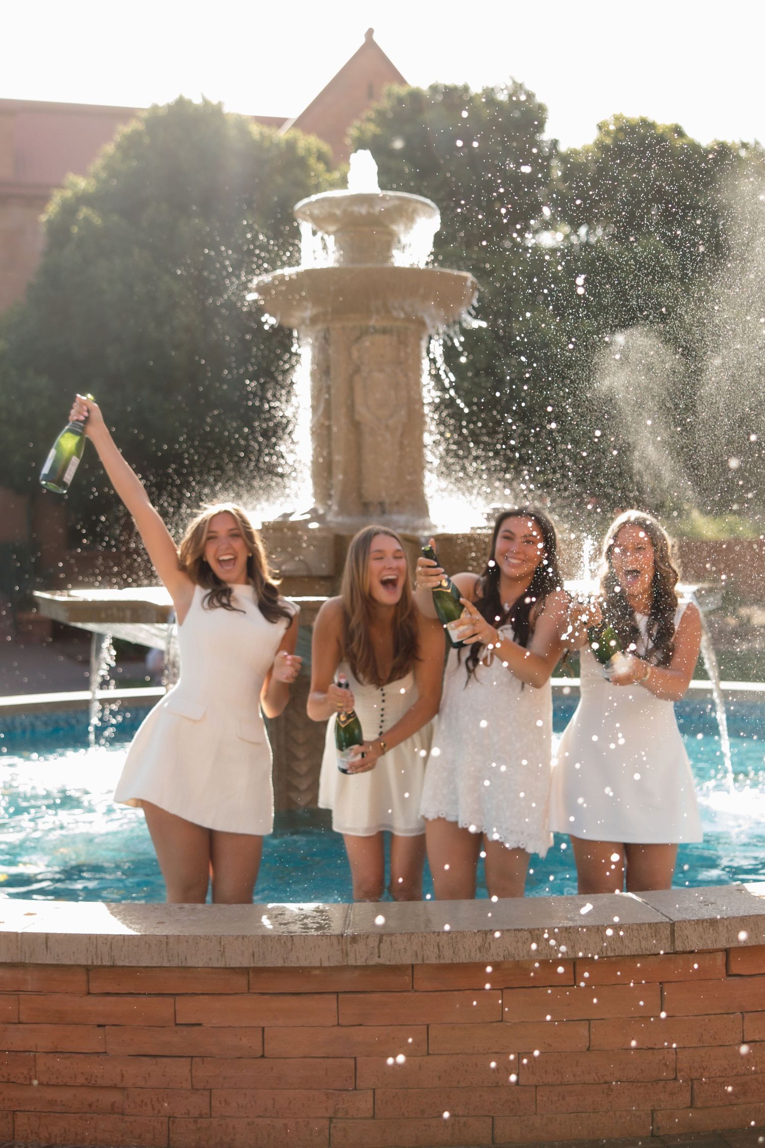 Four laughing women in white dresses pop champagne bottles at a campus fountain celebration.