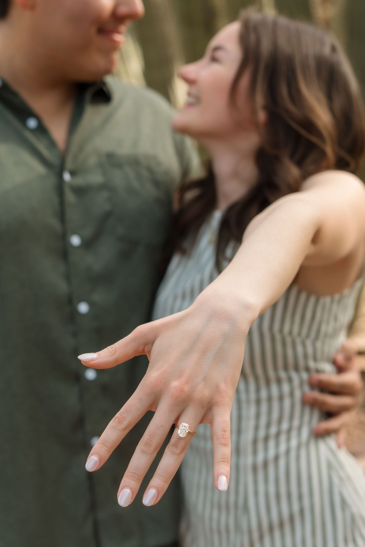Woman showing off engagement ring while embracing her fiance outdoors during engagement photo session.