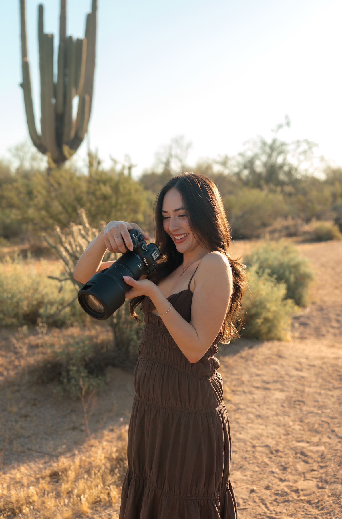 Woman in brown dress holds DSLR camera in desert landscape with saguaro cactus, golden hour lighting.