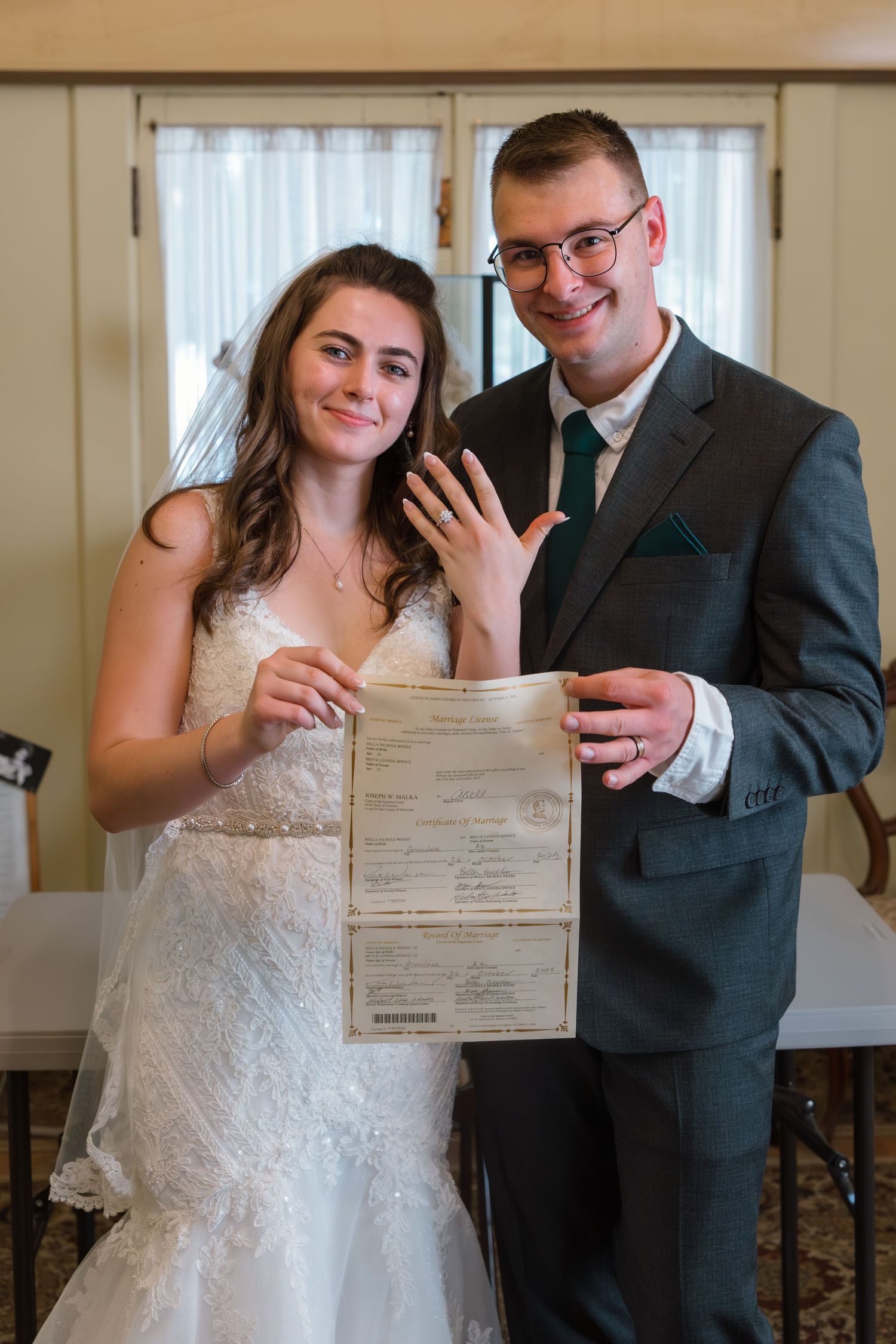 Smiling newlyweds holding marriage certificate and showing wedding rings at indoor wedding venue.
