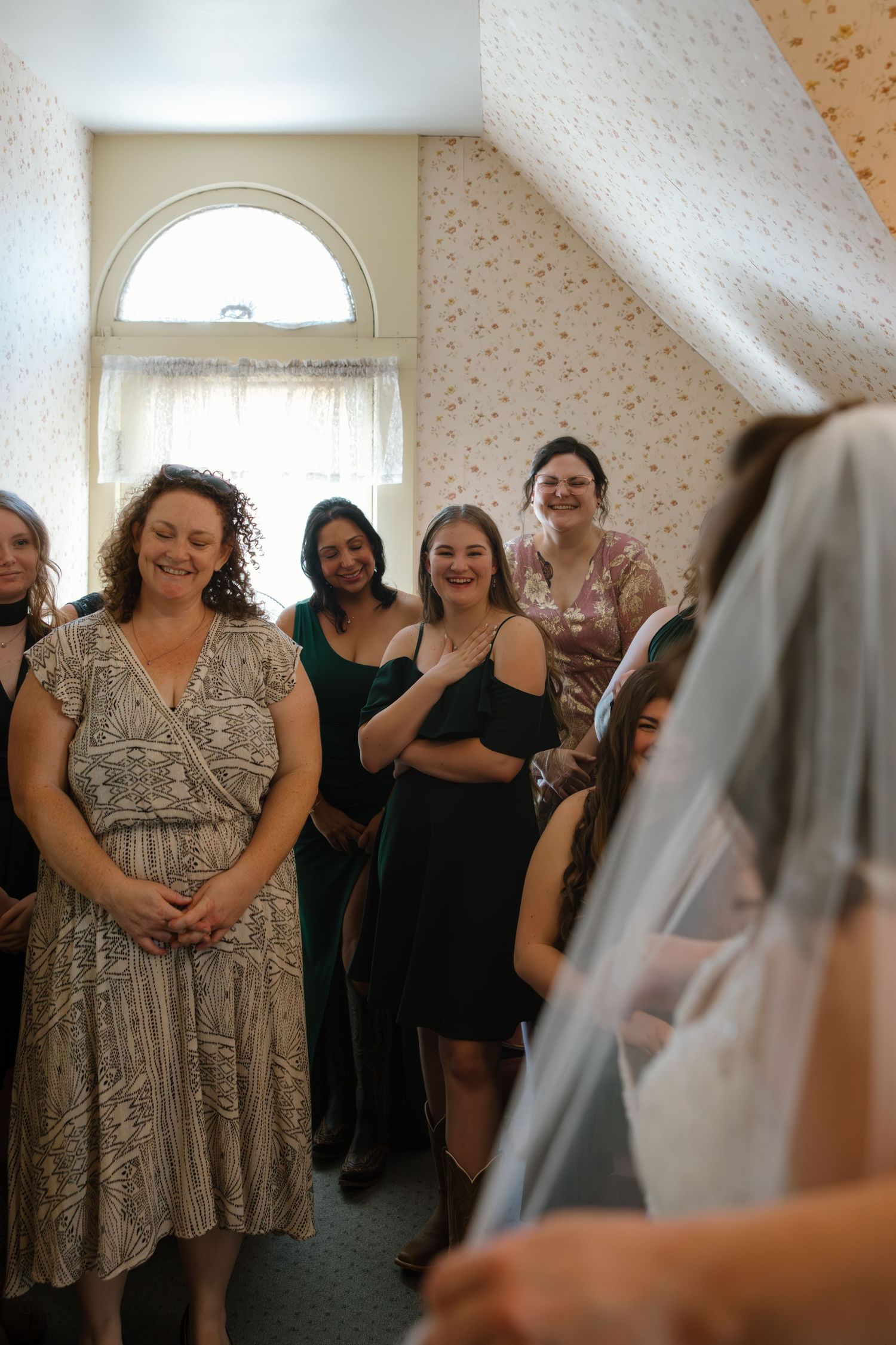 Bridesmaids and guests smile with joy as the bride in a veil reveals her wedding look in a decorated room.