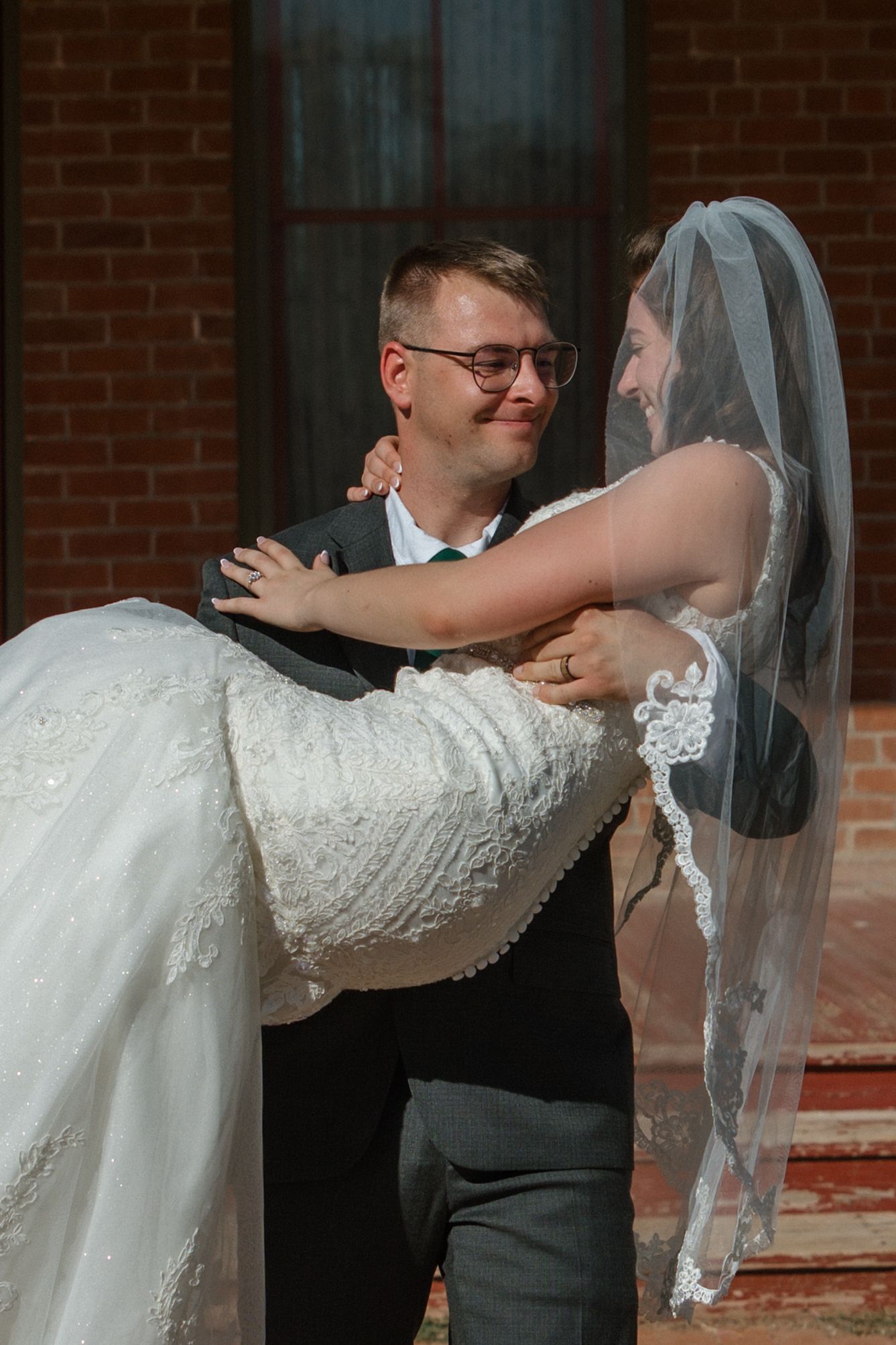Smiling groom in gray suit carries his laughing bride in a lace wedding dress outside a brick building.