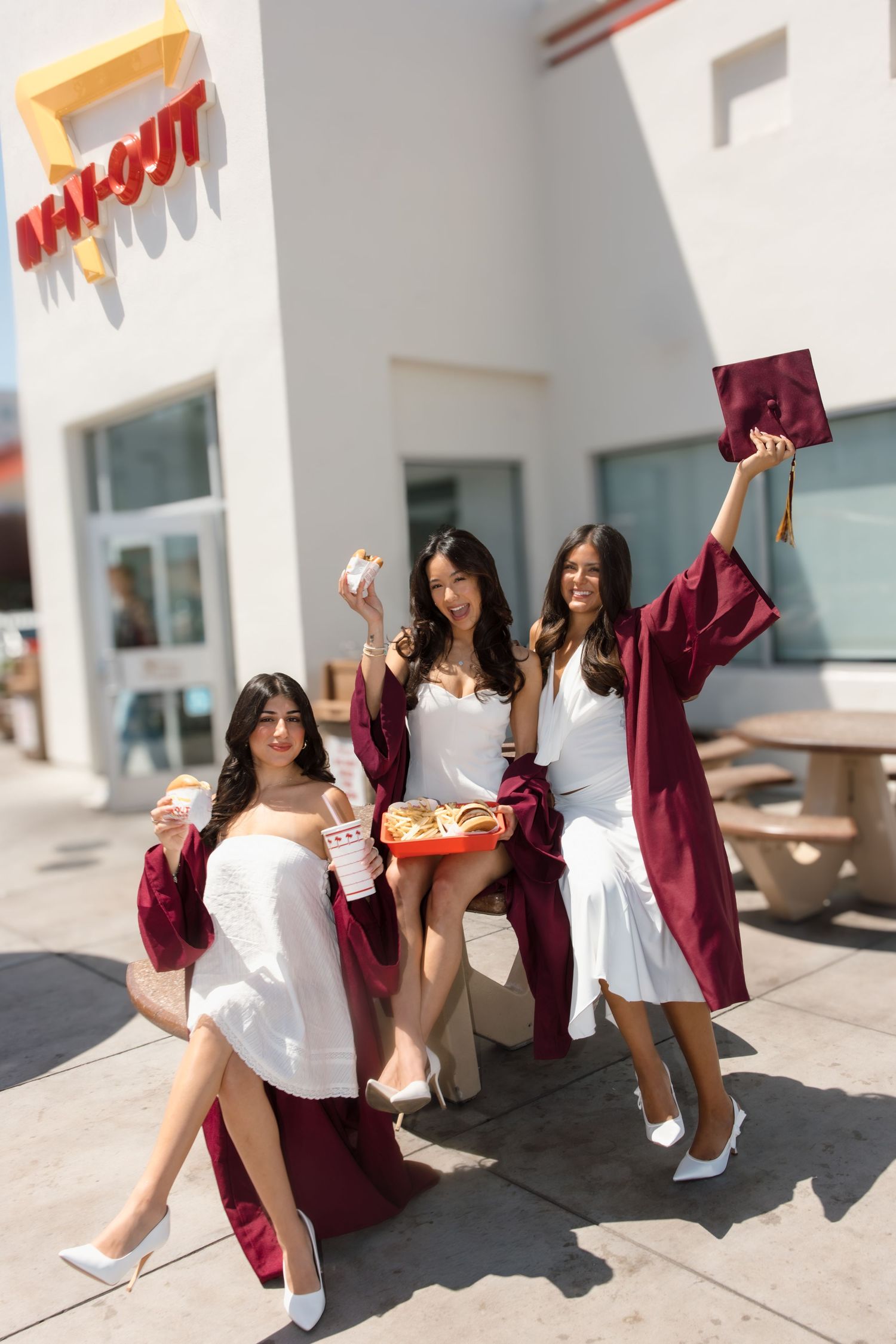 Three smiling female graduates in burgundy robes enjoy In-N-Out Burger food and drinks outside the restaurant.