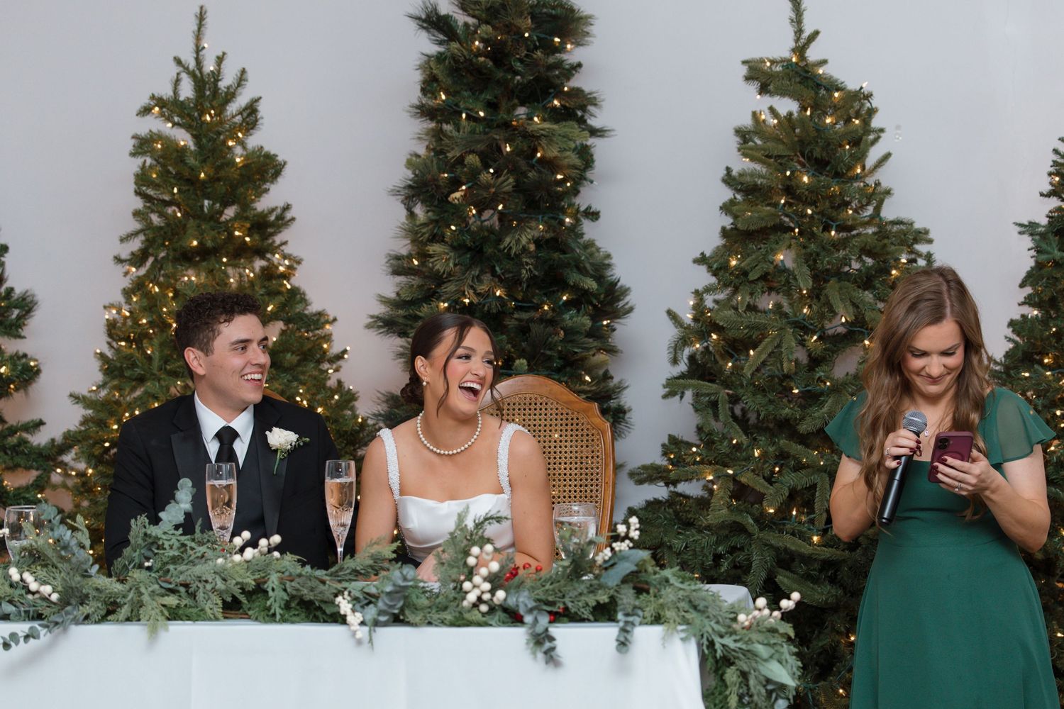 Bride laughing at sweetheart table as bridesmaid in green dress gives a wedding toast with Christmas trees behind.