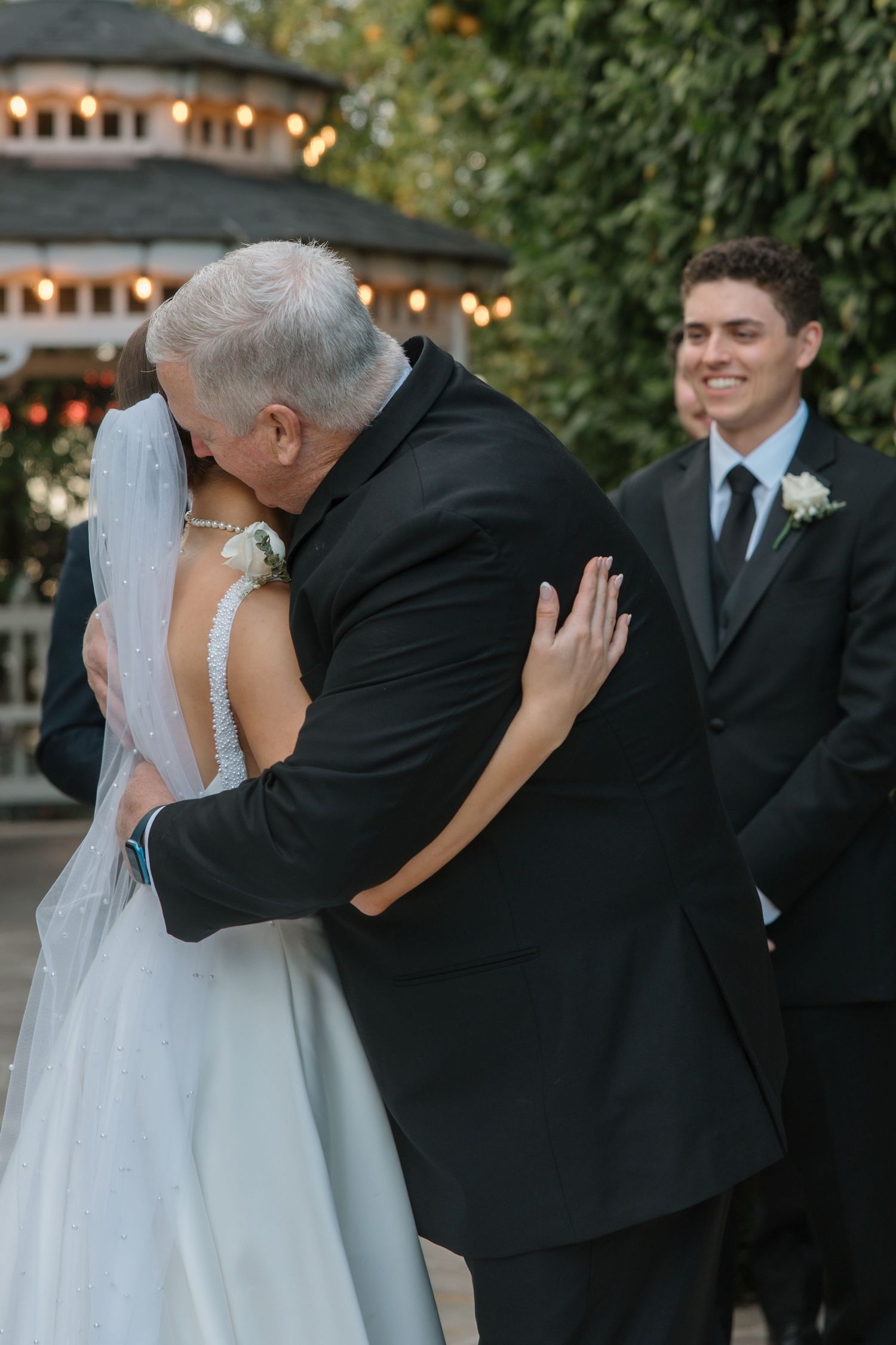 Father of bride shares emotional hug with daughter as smiling groom watches at outdoor ceremony.