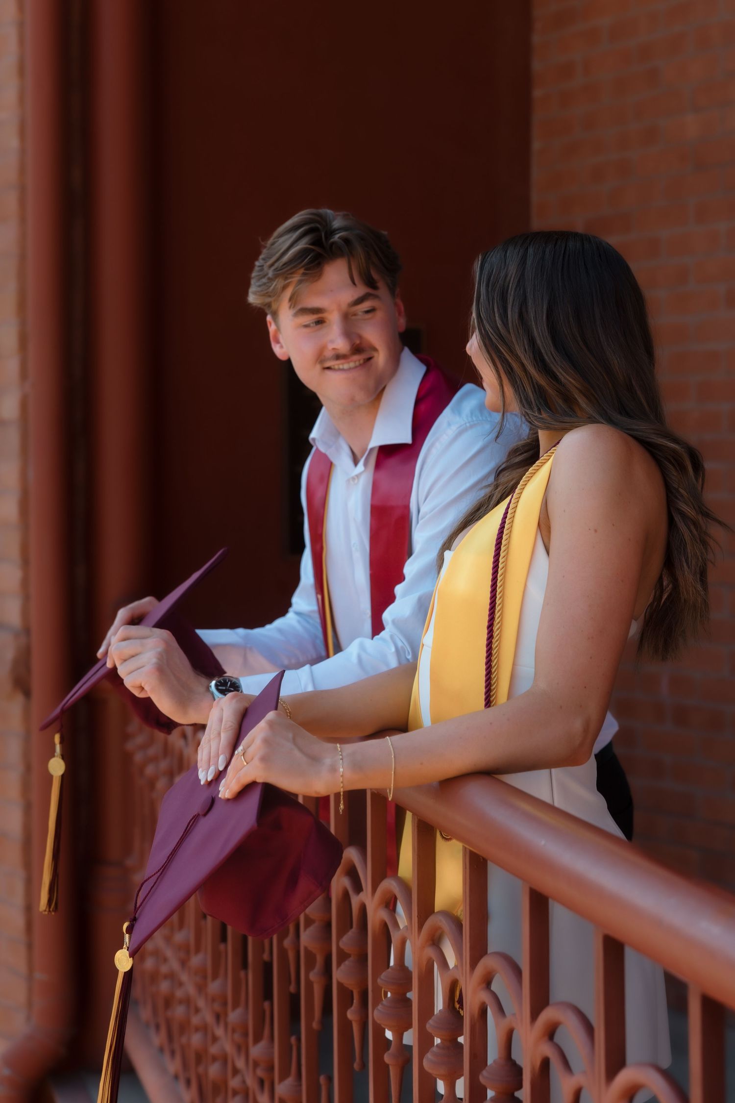 Graduate smiles at woman in white dress while leaning on decorative iron railing on campus.