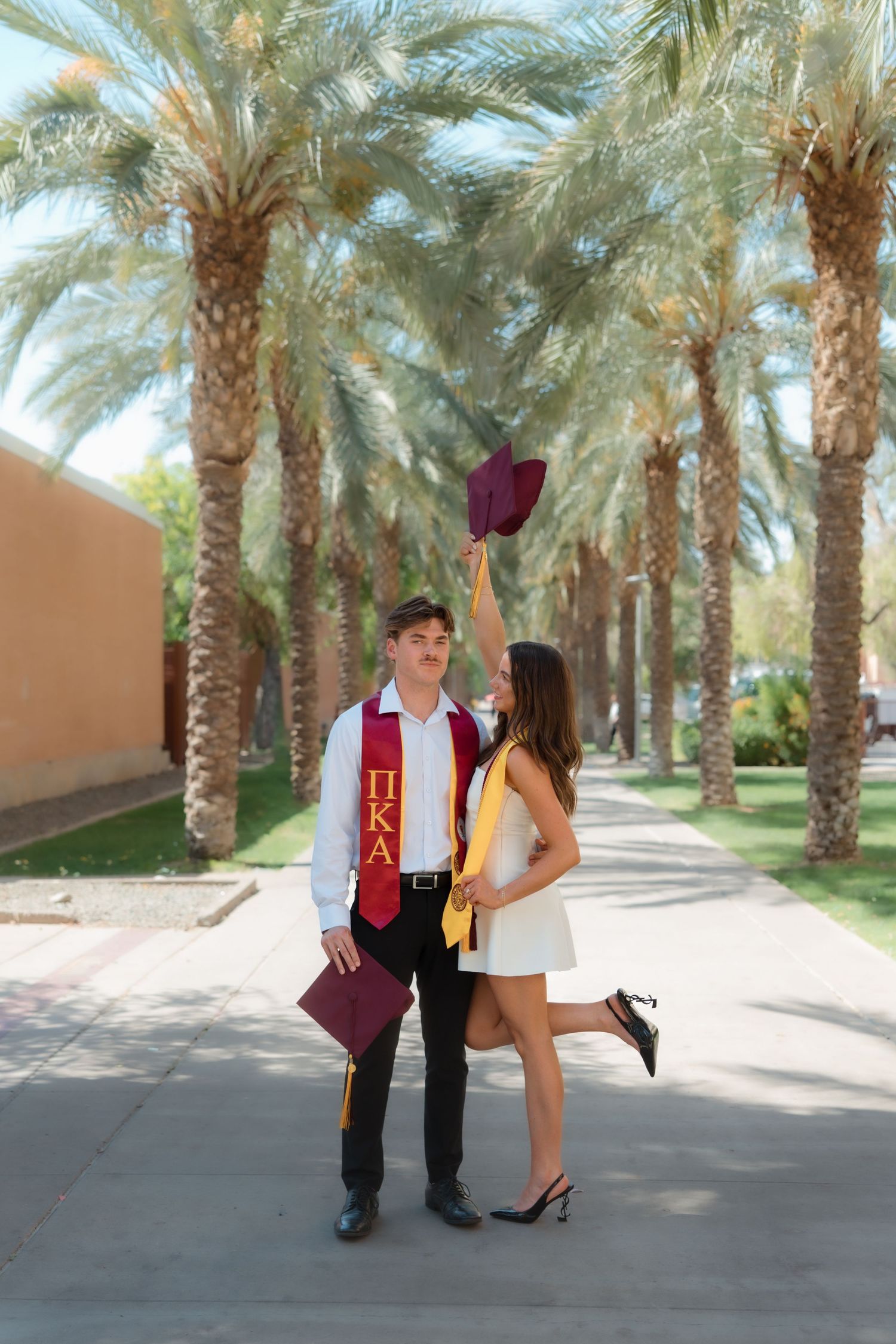 Graduating couple on palm-lined path, woman raising maroon cap while man holds one.