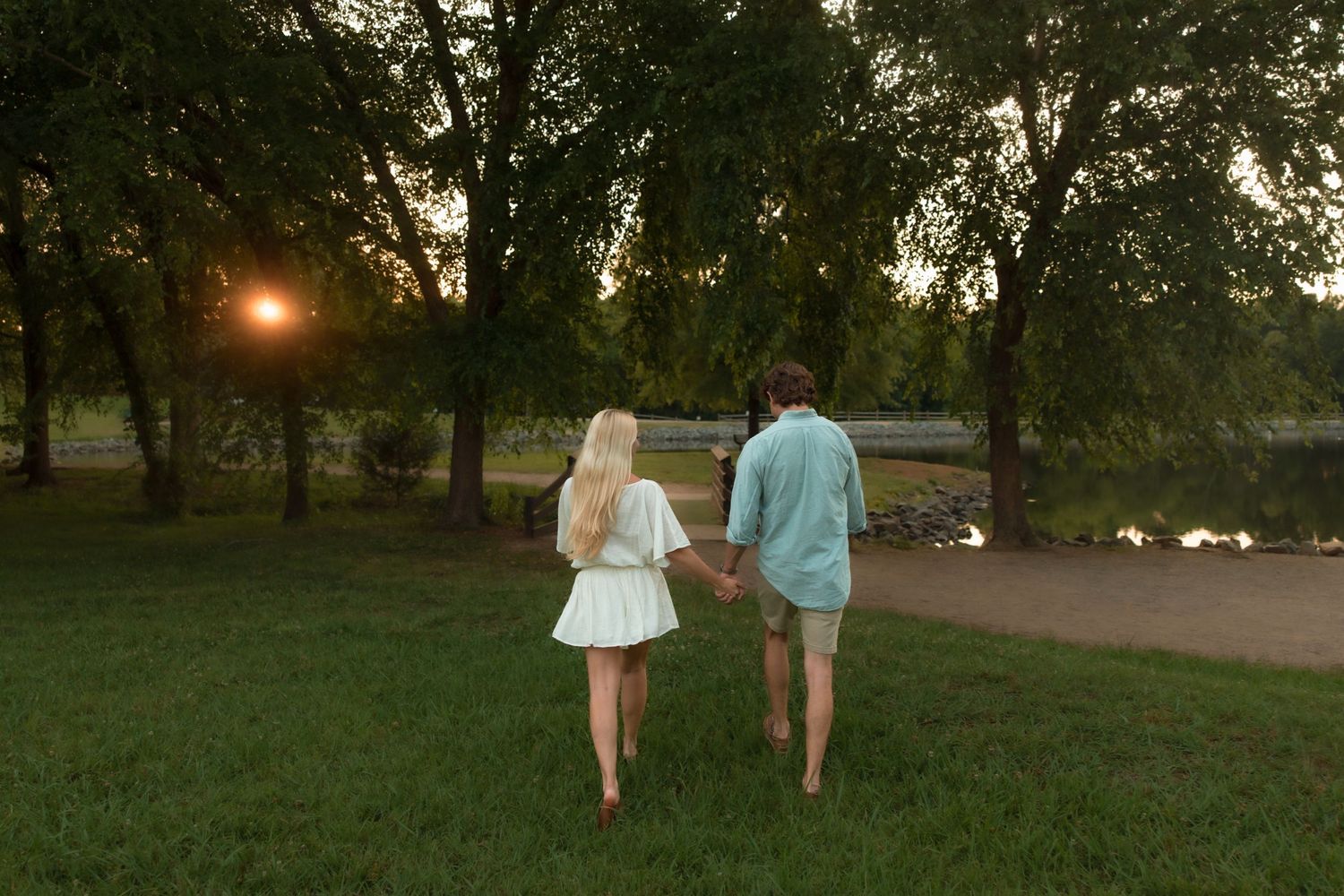 A couple walks hand in hand barefoot through a park at sunset, viewed from behind.