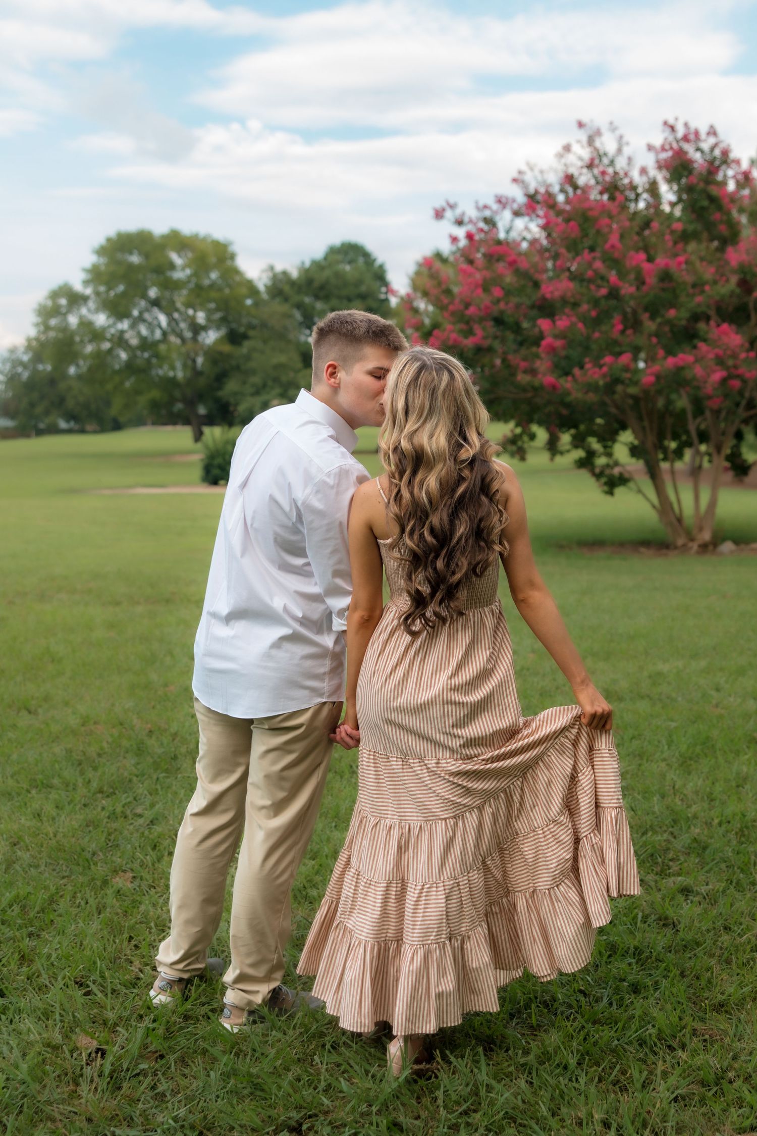 Couple sharing a kiss outdoors, woman in flowing blush maxi dress holding skirt, man in white shirt, pink flowering tree behind.