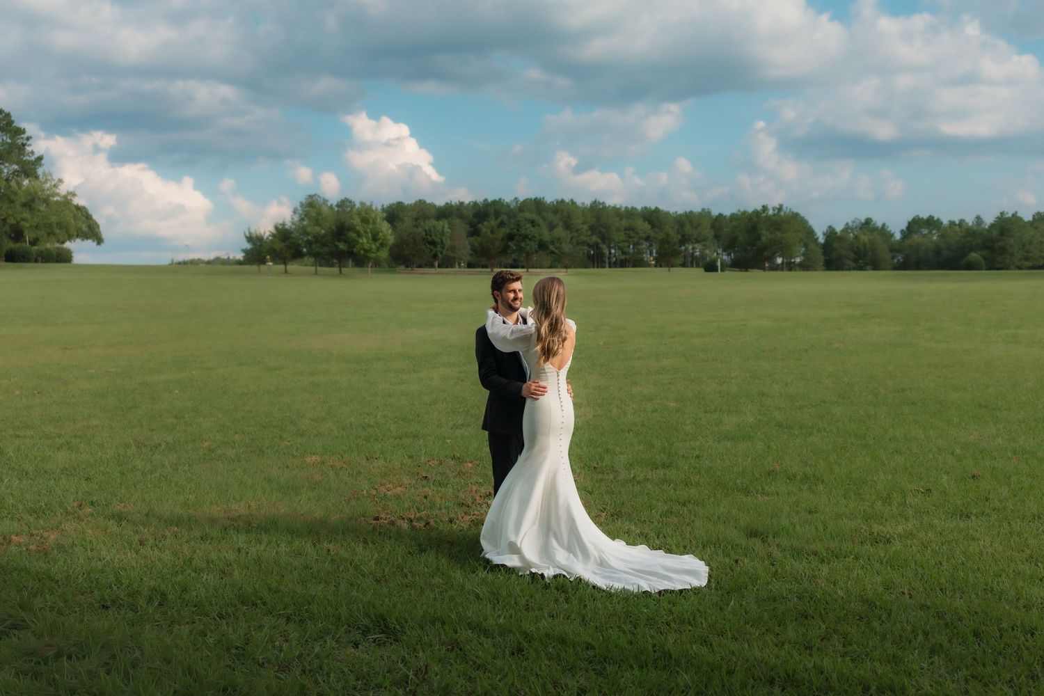 A bride and groom share a romantic moment in a lush green field under a blue sky on their wedding day.