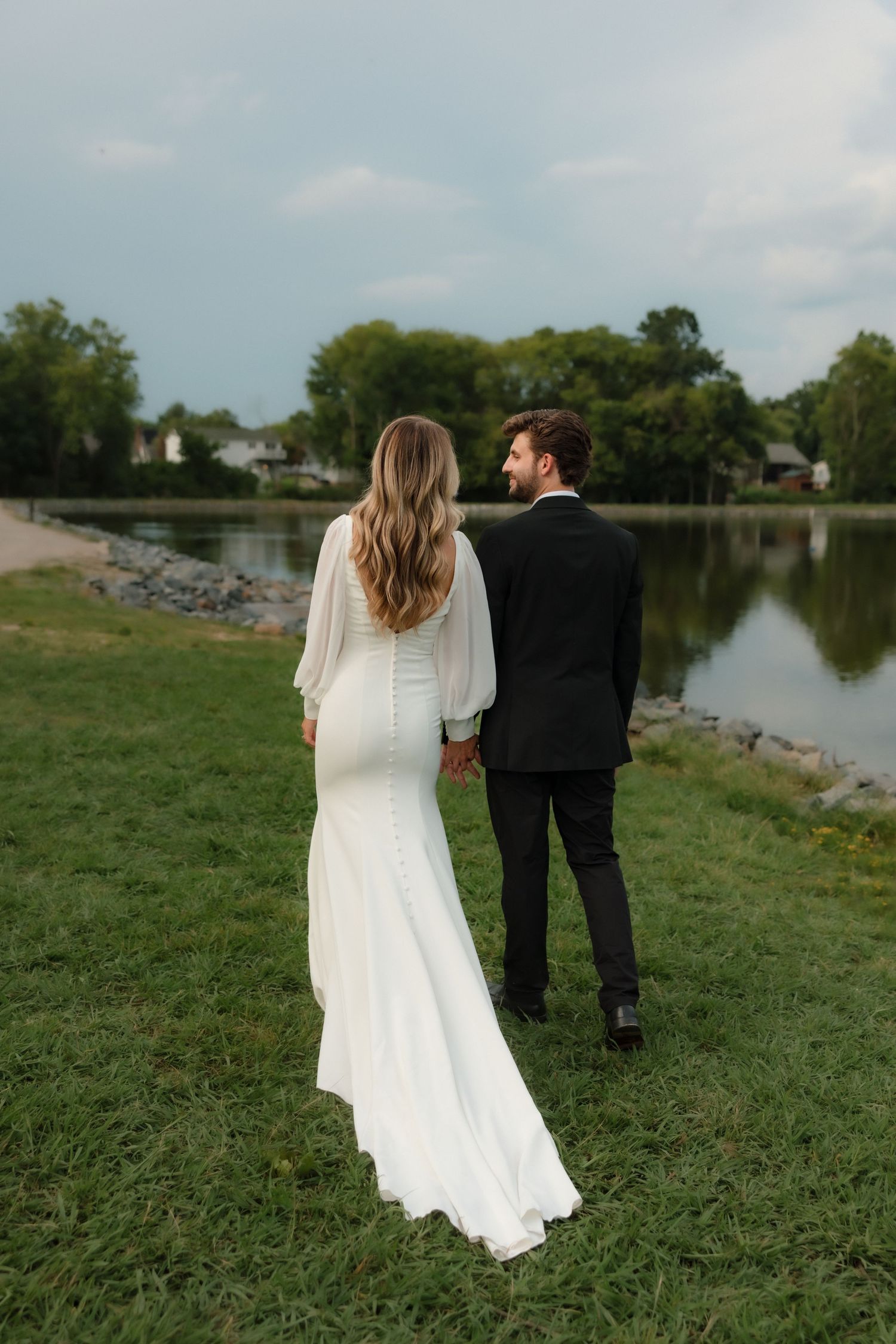 Bride and groom holding hands from behind, standing by a calm lakeside in wedding attire.
