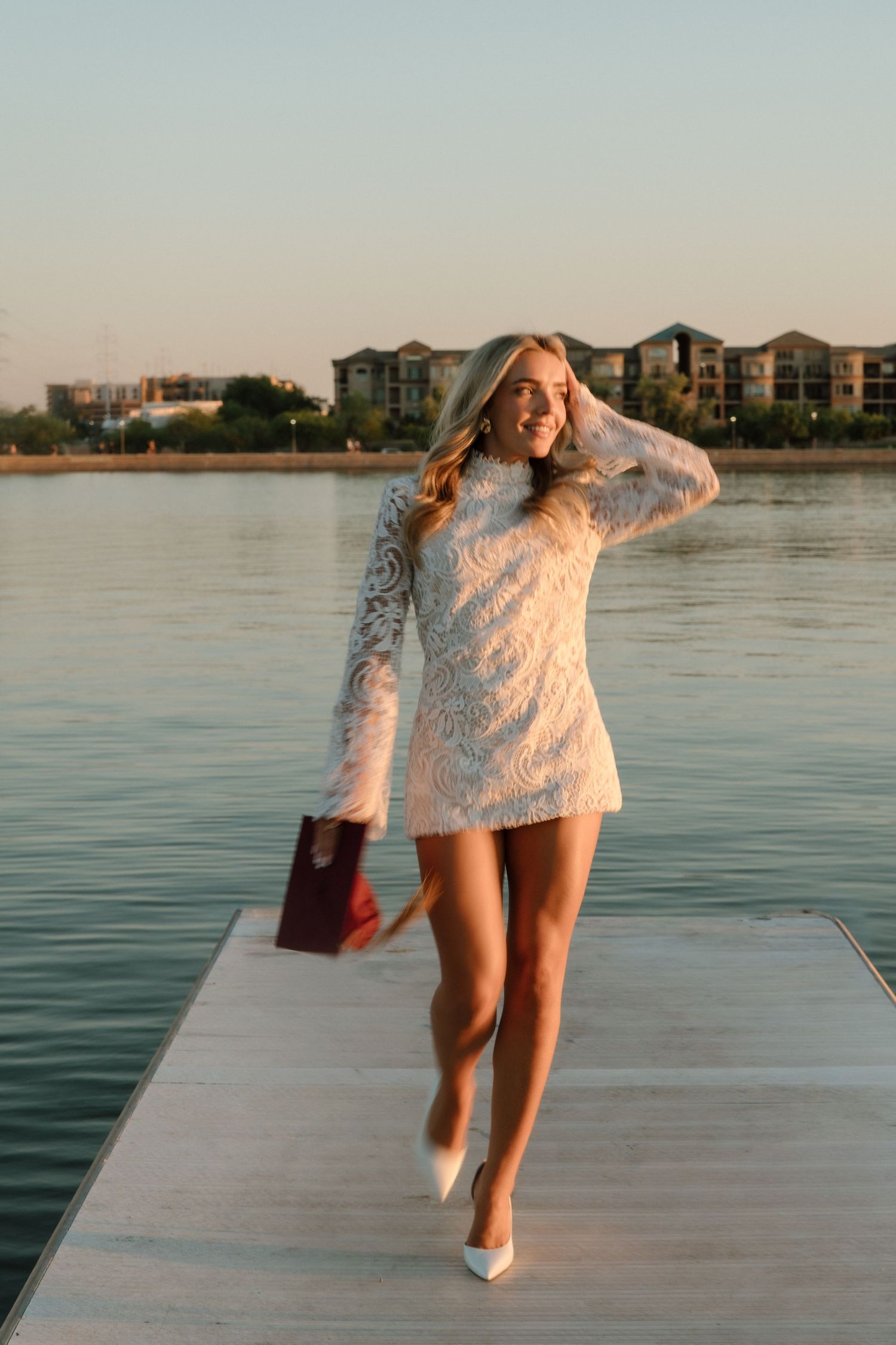Woman in white lace mini dress and heels poses on dock by waterfront at sunset.