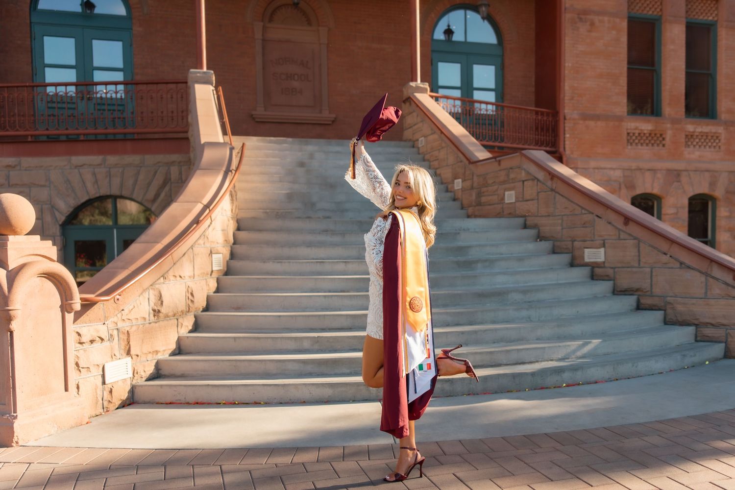Graduate in white gown waves maroon cap on steps of ASU campus building in warm golden sunlight.
