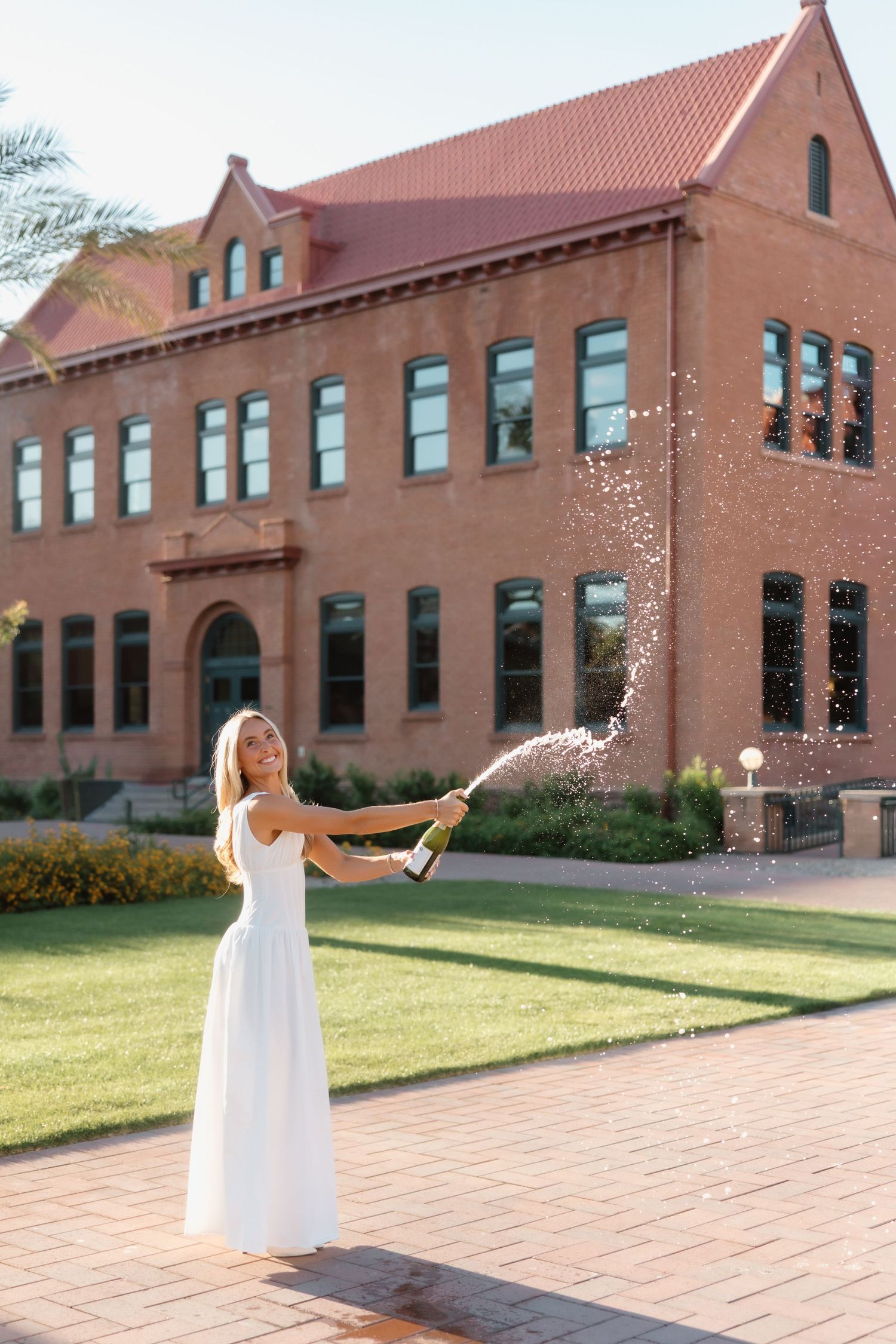 Woman in white dress popping champagne bottle outdoors near red brick university building on sunny day.