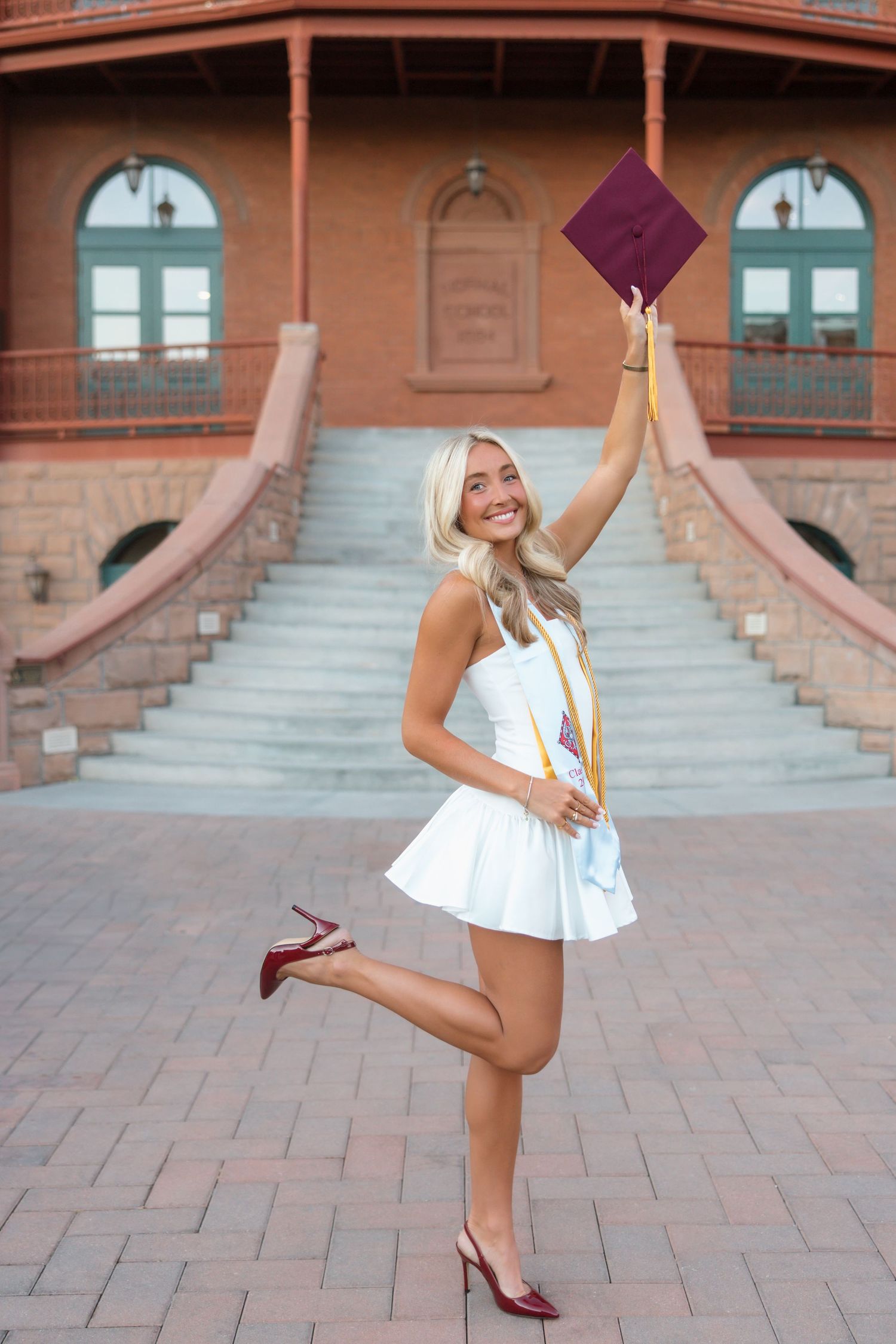 Smiling blonde graduate in white dress tosses maroon cap outside brick university building with stairs.