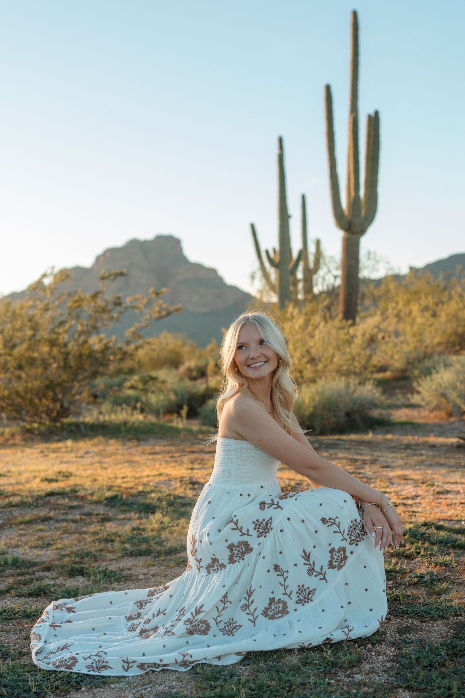 Smiling blonde woman in white floral dress sitting in Arizona desert with saguaro cactus and mountains at golden hour.
