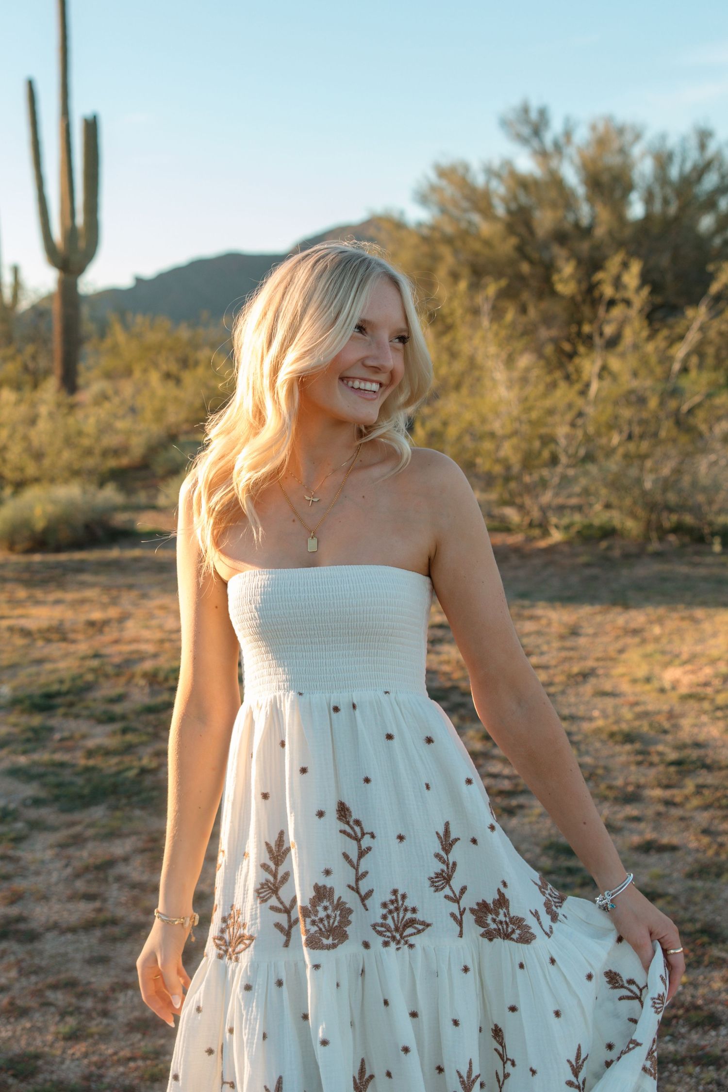 Smiling blonde woman in white floral strapless maxi dress posing in desert landscape with saguaro cactus at golden hour.