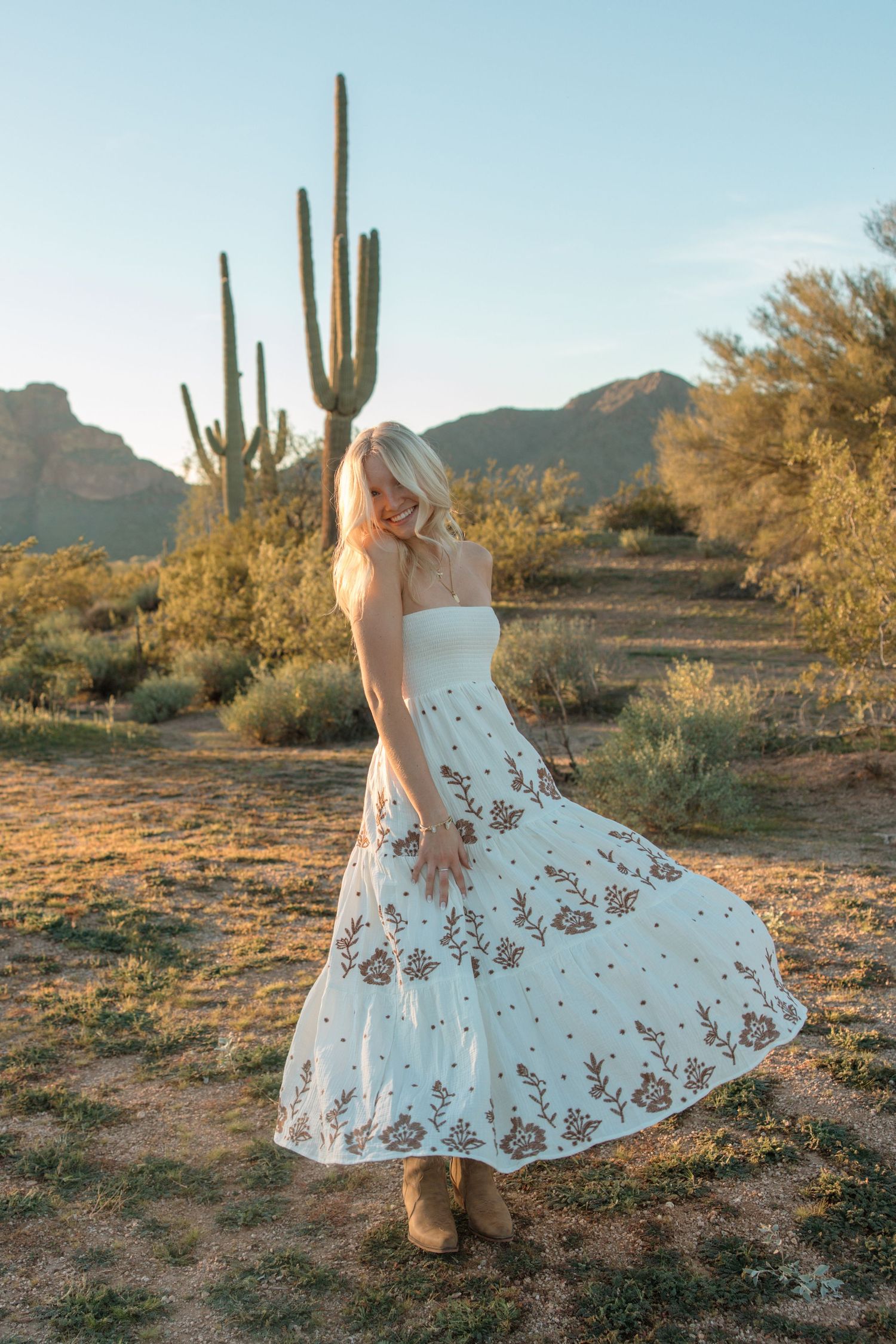 Blonde woman in white floral maxi dress posing in Arizona desert with saguaro cactus and mountains at golden hour.
