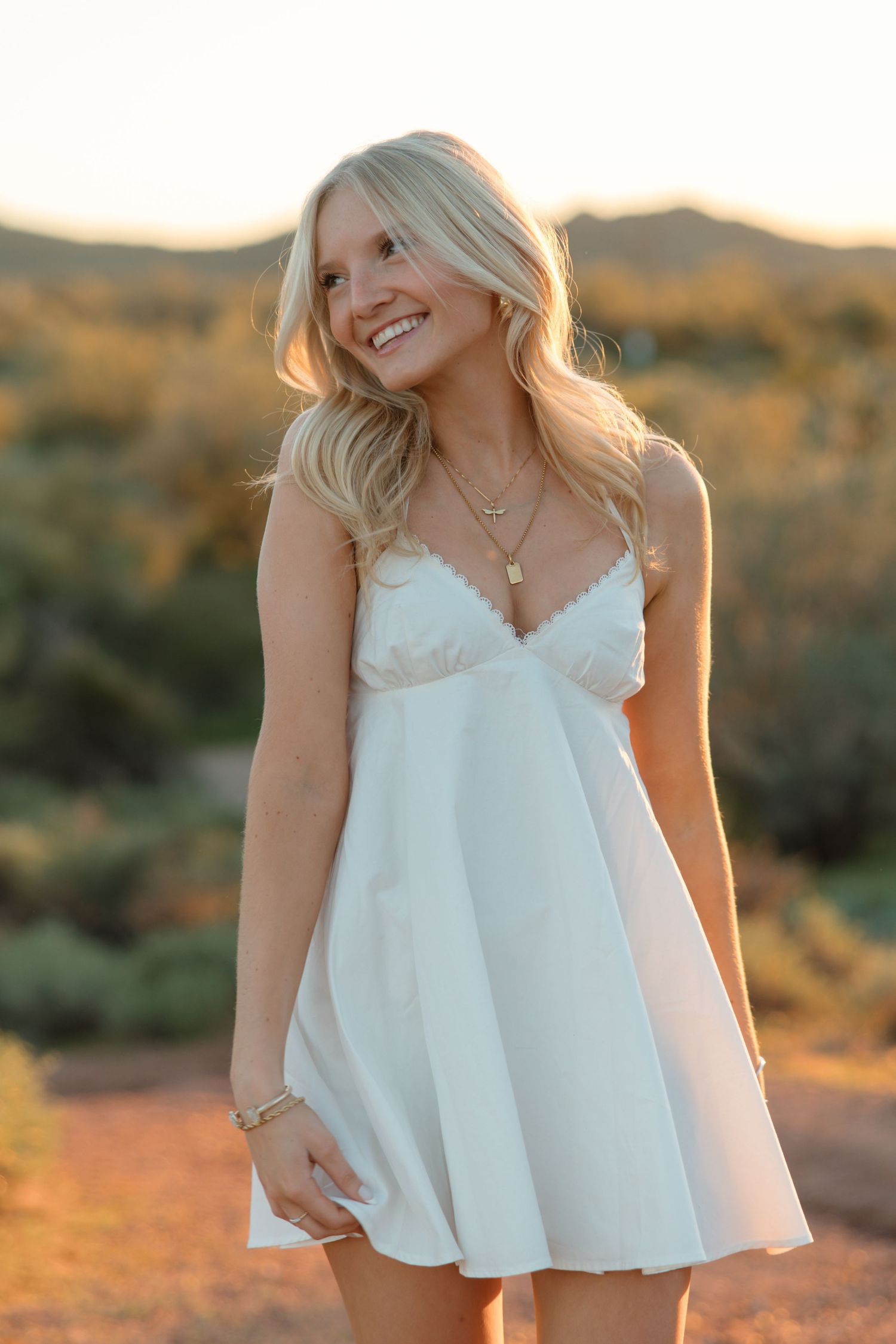 Smiling blonde woman in white mini dress posing outdoors at golden hour with desert landscape background.