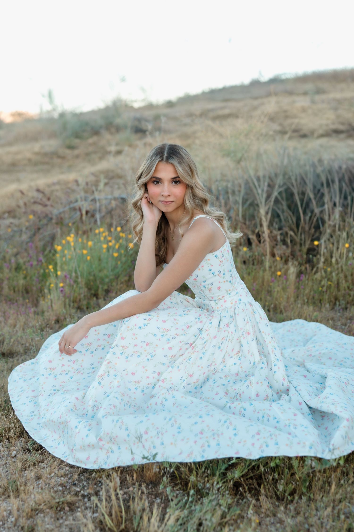 Young woman in a floral white maxi dress sitting gracefully in a golden field during sunset.