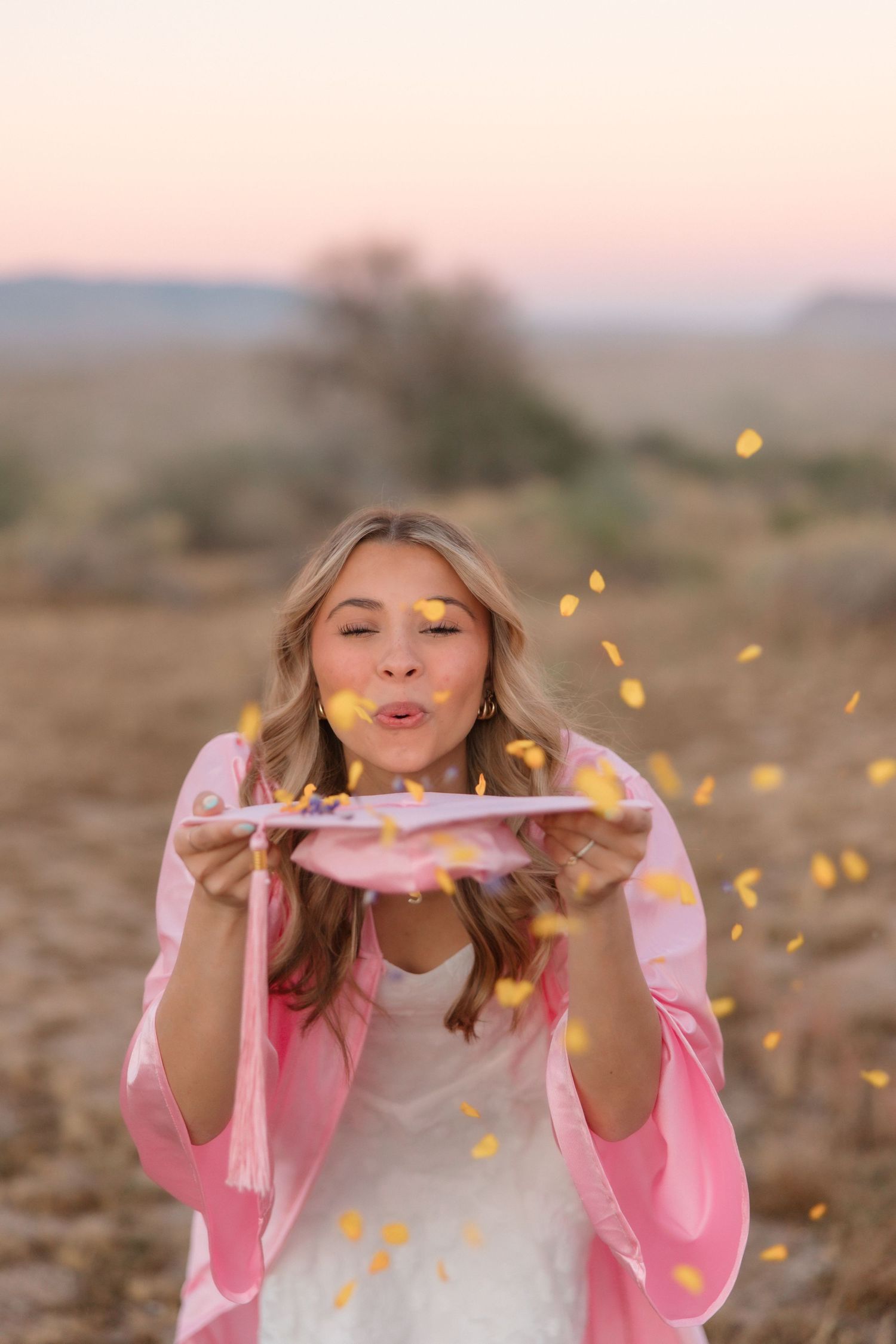 Young woman in pink jacket blowing yellow confetti from a plate outdoors at sunset in a golden field.