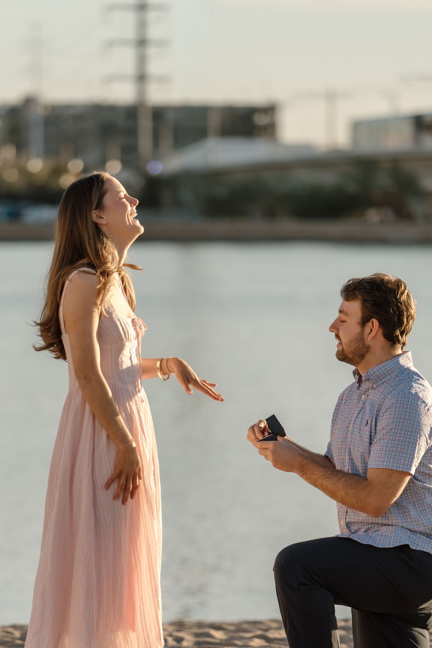 Joyful woman reacting to marriage proposal from kneeling man holding ring box by river.