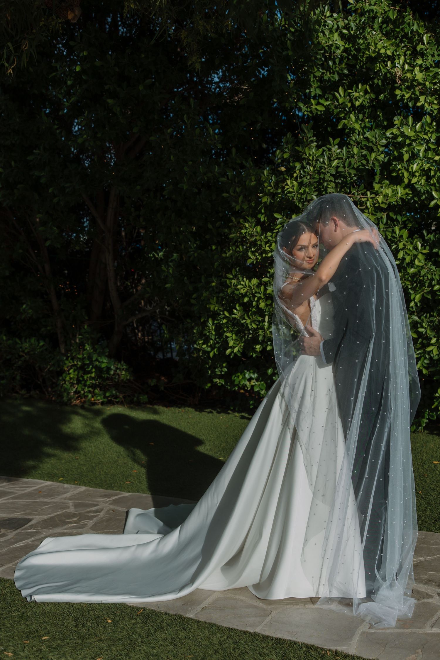 Bride and groom embracing under a long pearl veil with a lush green garden backdrop on wedding day.