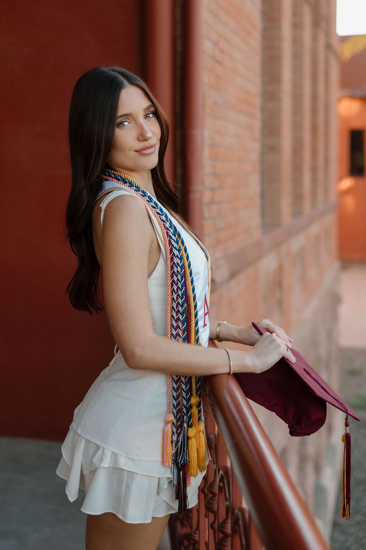 Female graduate in white dress holds maroon cap while leaning on iron railing of red brick campus building.