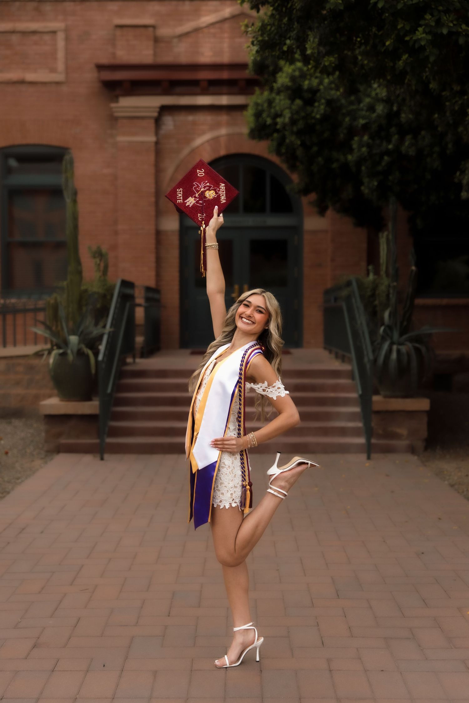ASU graduate in white dress posing with one leg raised, holding maroon graduation cap aloft in front of historic brick building.