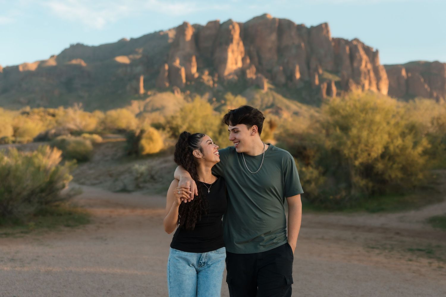 A smiling couple gazes at each other with their arms around each other in a scenic desert landscape.