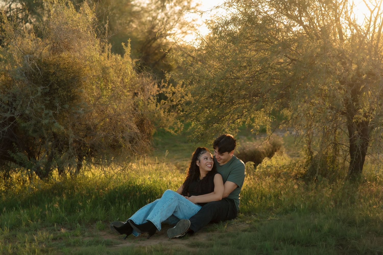 A couple sits together in a sunlit meadow surrounded by lush green trees during golden hour.