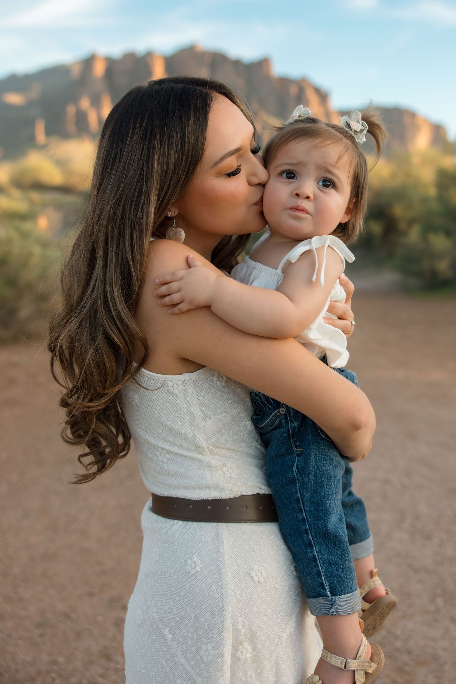 Mother in white lace dress kissing her toddler daughter on the cheek with rocky mountain background.
