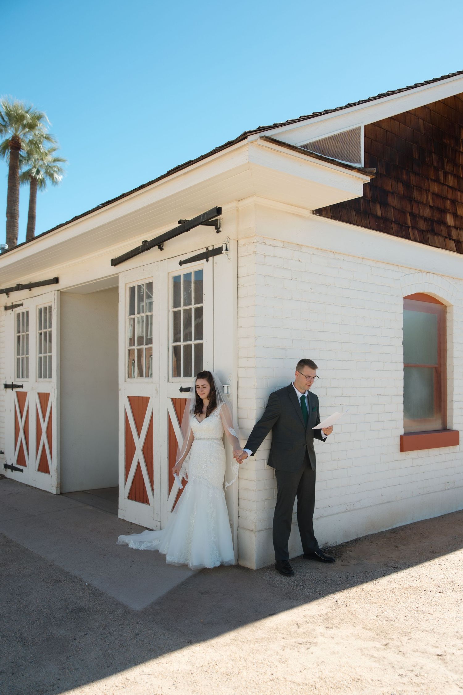 Bride and groom hold hands outside rustic white barn venue with palm trees under clear blue sky.
