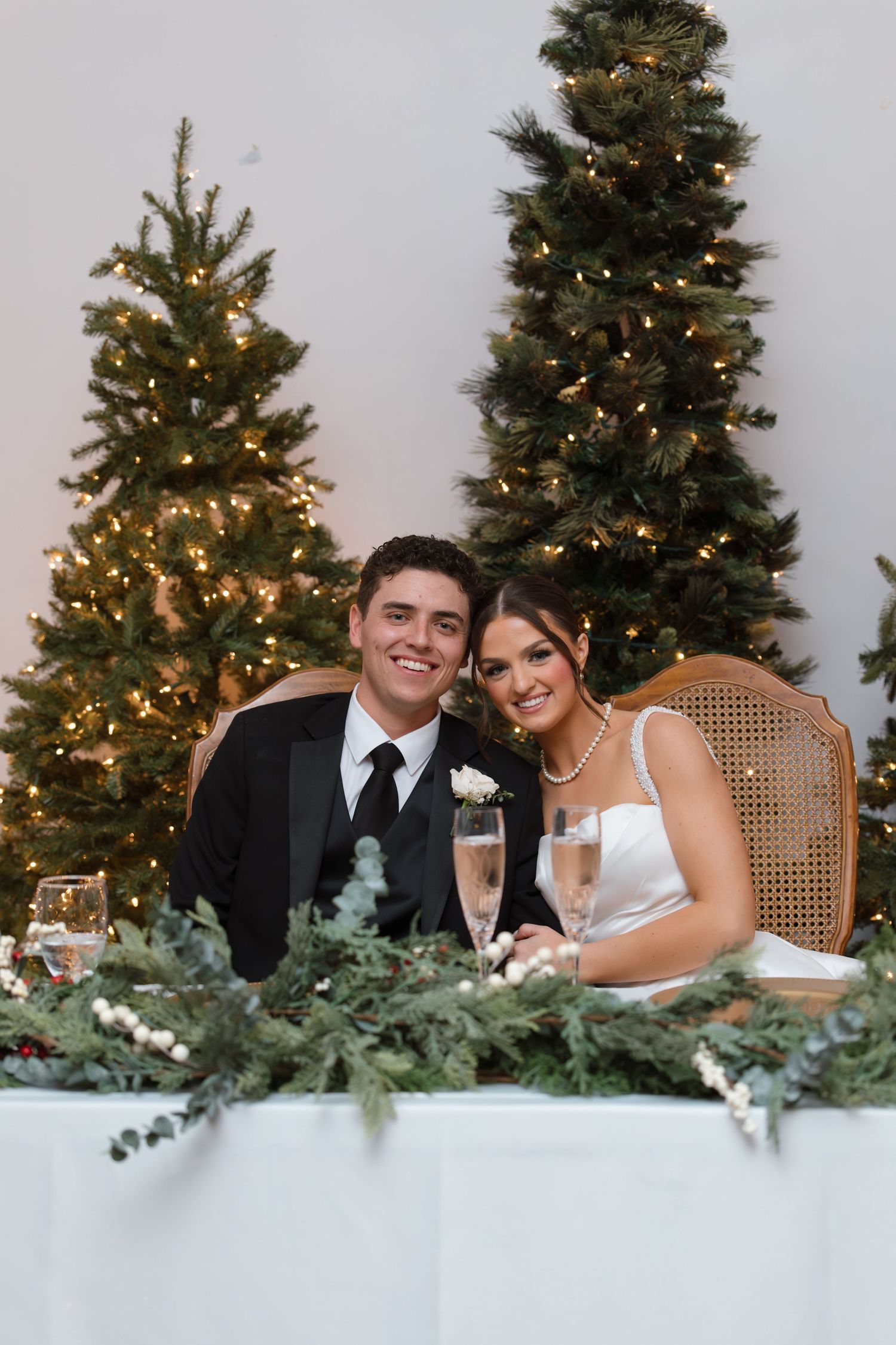 Smiling bride and groom seated at sweetheart table with champagne flutes and lit Christmas trees behind them.