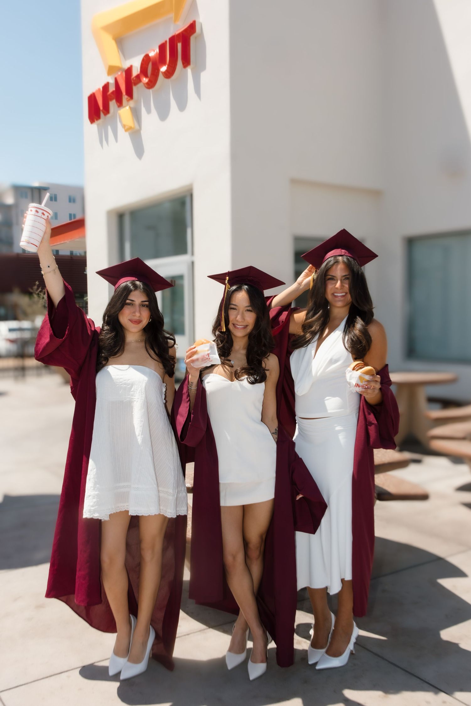 Three female graduates in burgundy robes and white dresses celebrate outside In-N-Out Burger holding drinks.