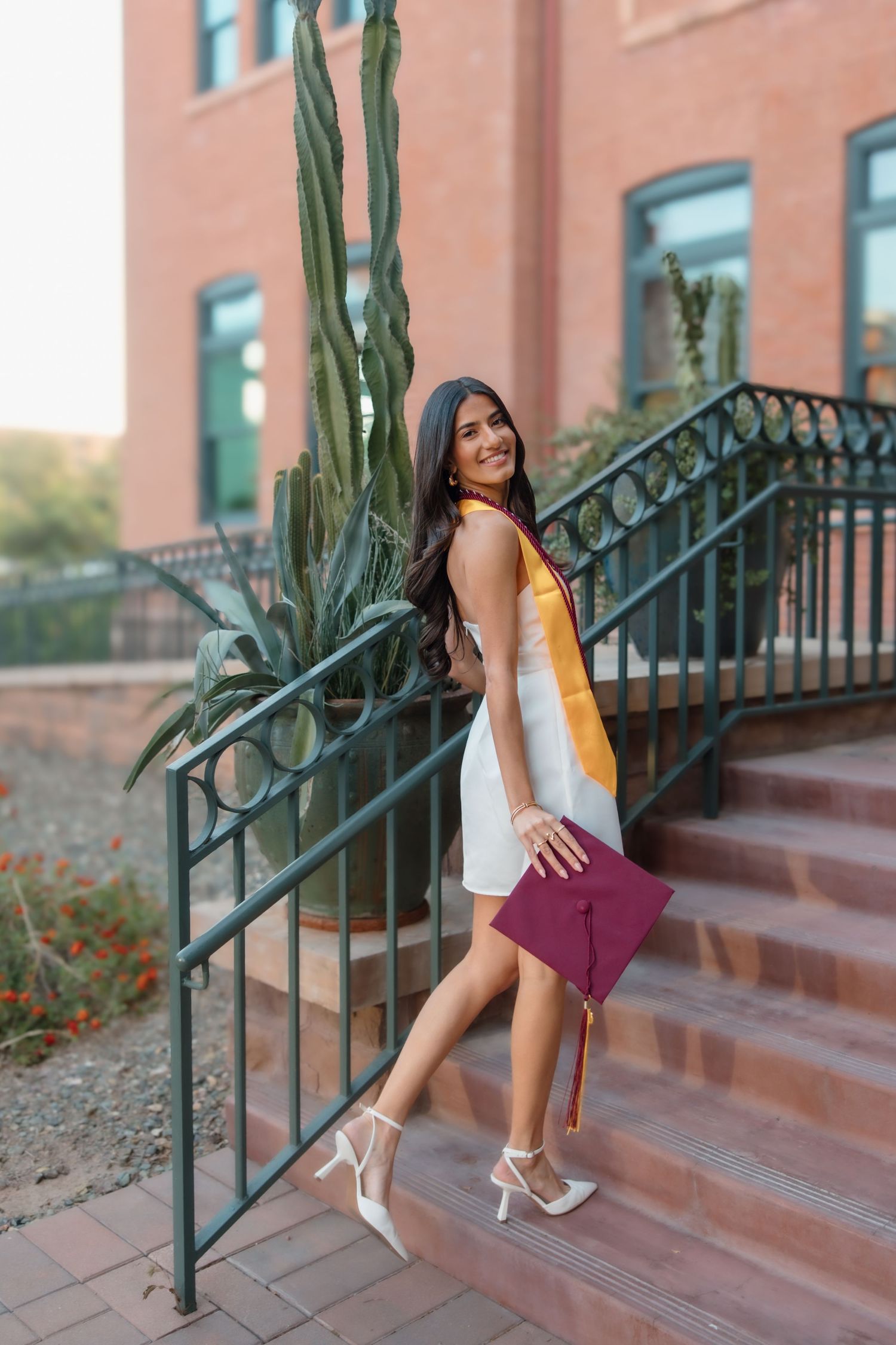 Young woman in white dress holding maroon graduation cap on university steps with cactus backdrop.