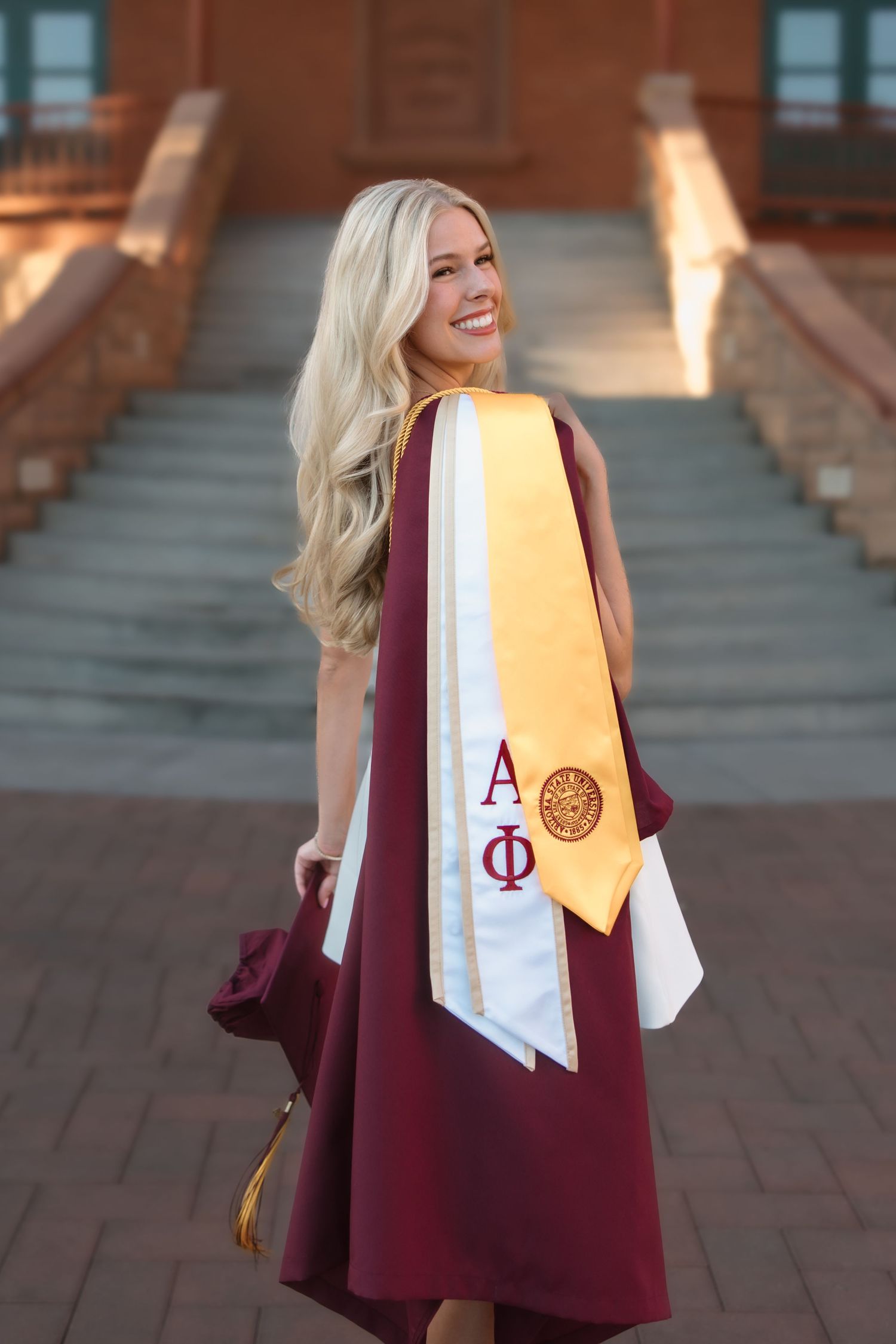 Smiling blonde graduate in maroon gown with Alpha Phi sash poses on sunlit campus steps.