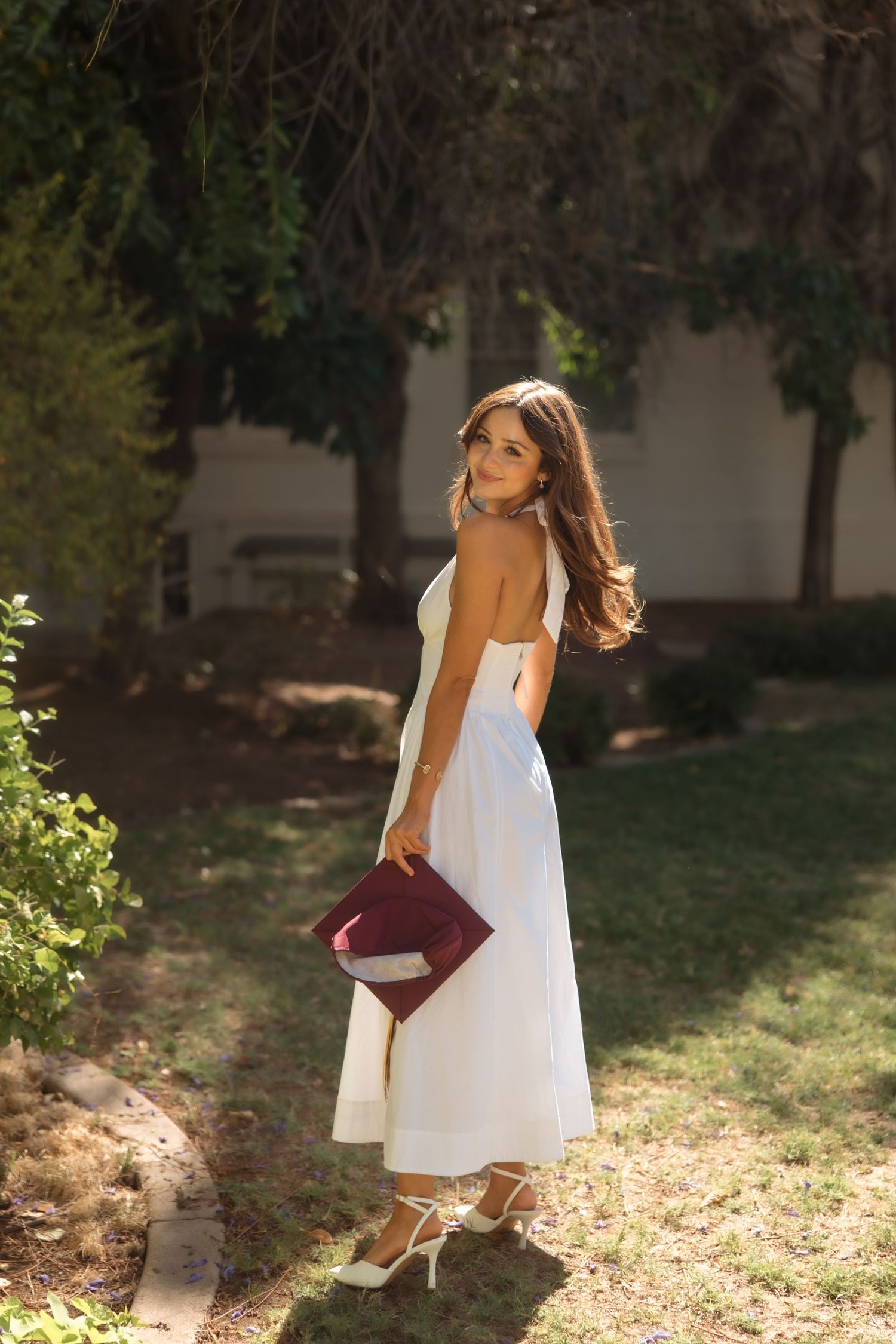 Young woman in white midi dress holding burgundy clutch, posing outdoors in sunlit garden with lush green trees.