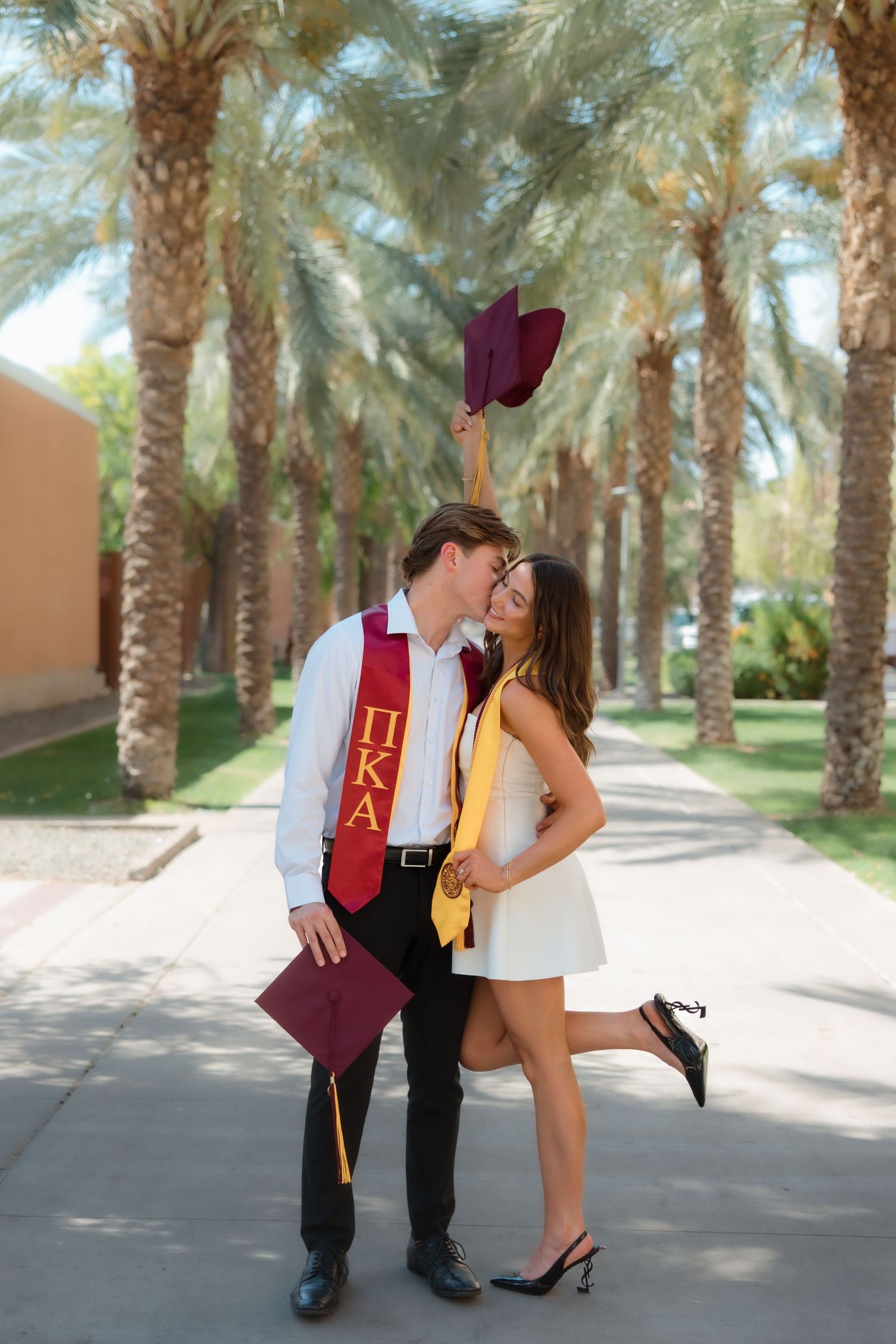 Graduating couple sharing a kiss on palm tree-lined campus path, cap raised high.