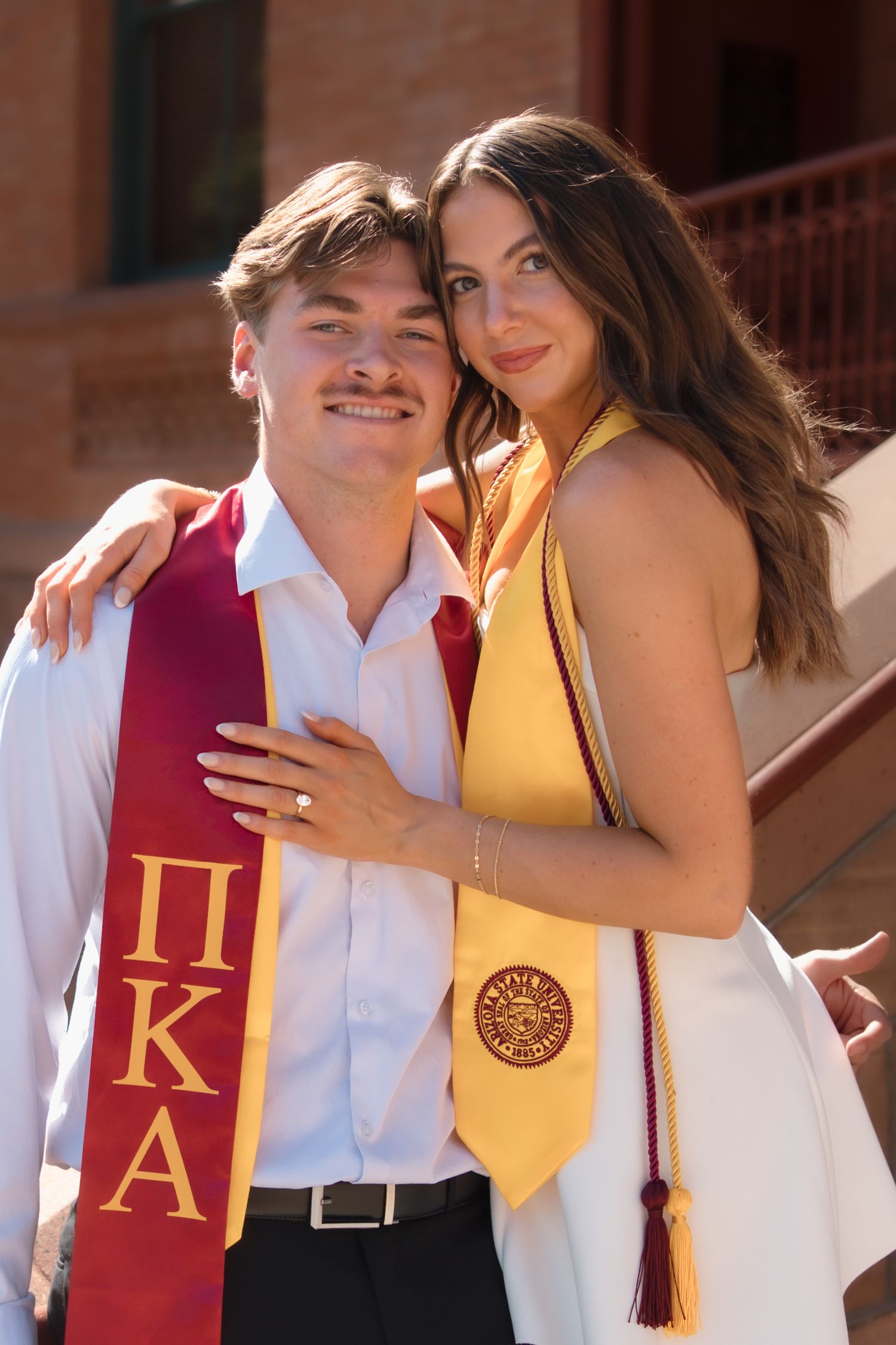 Two college graduates in maroon and gold honor stoles pose together smiling on campus stairs.