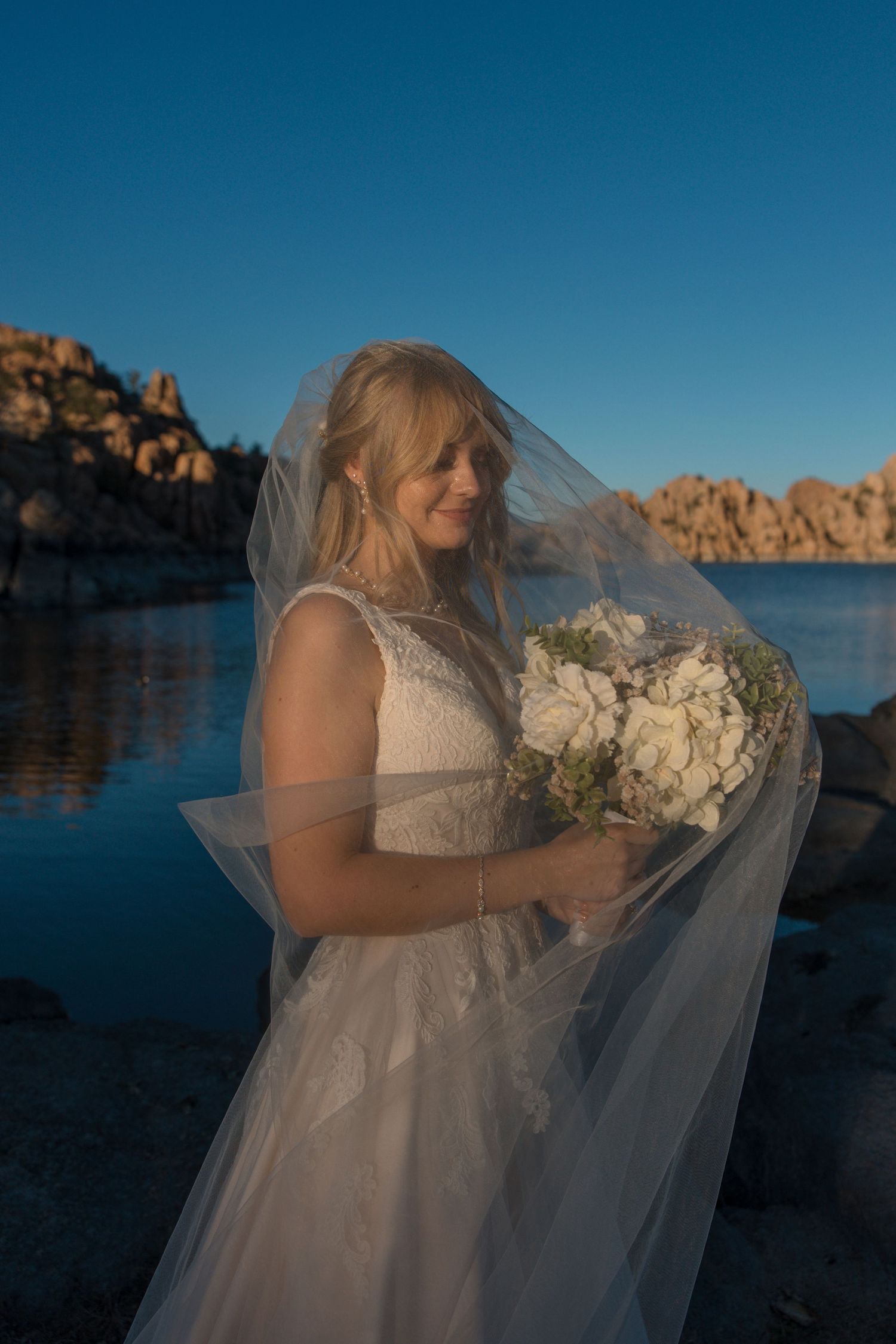 Beautiful bride holds white bouquet as veil drapes over her face near a scenic rocky lake at sunset.